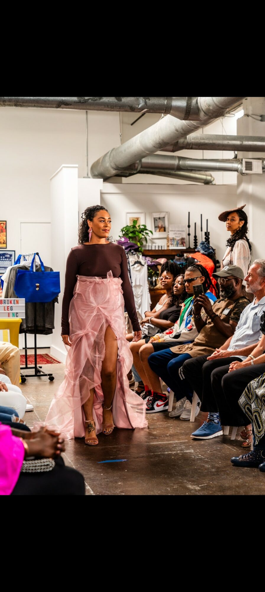 Person in pink skirt walking past seated group in industrial-style room with pipes and plants