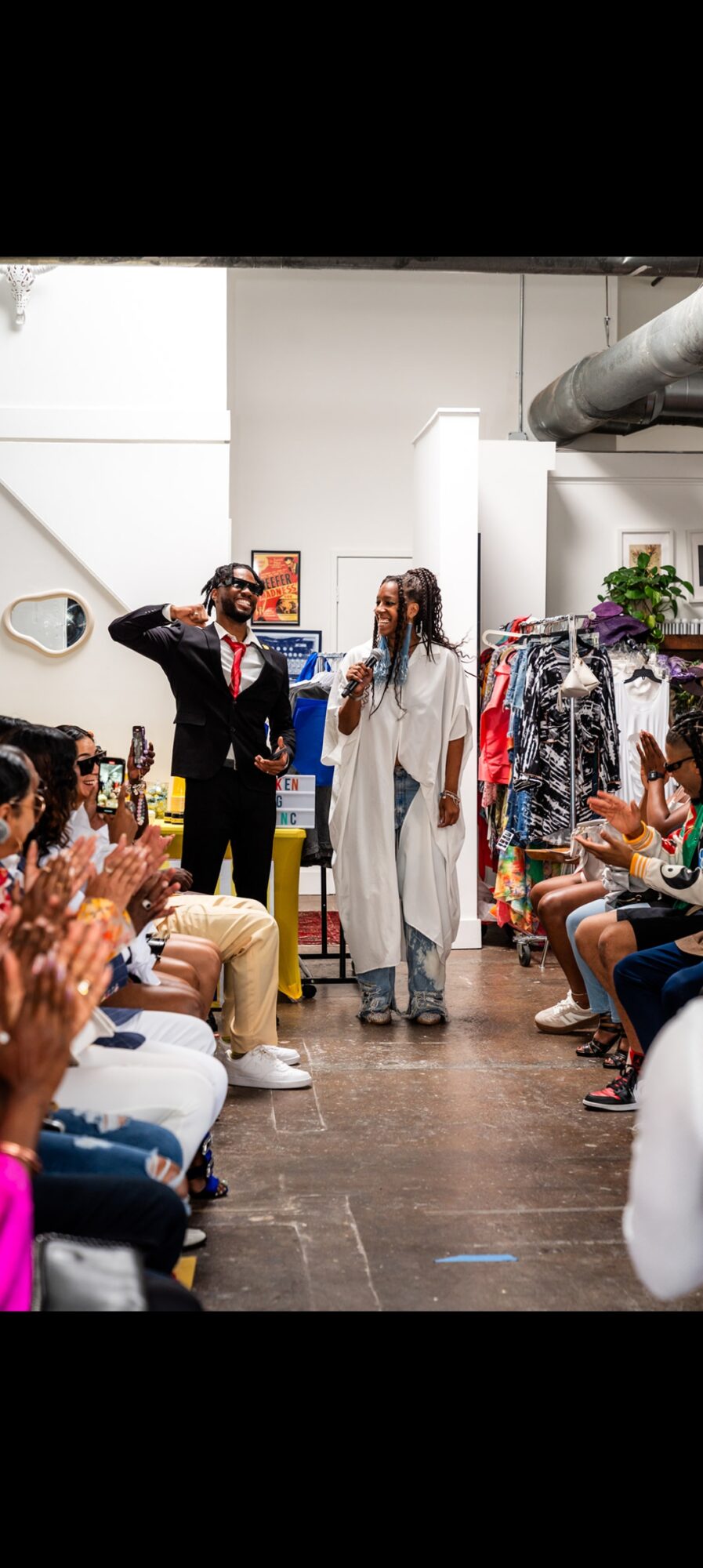 People seated on both sides of a runway, with two models walking and posing, in a fashion show setting.