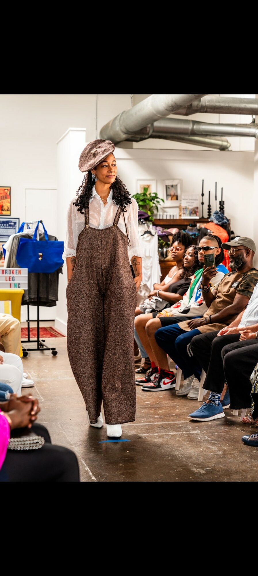 Person walking down runway in brown overalls, white shirt, and hat, audience seated on both sides, industrial background.