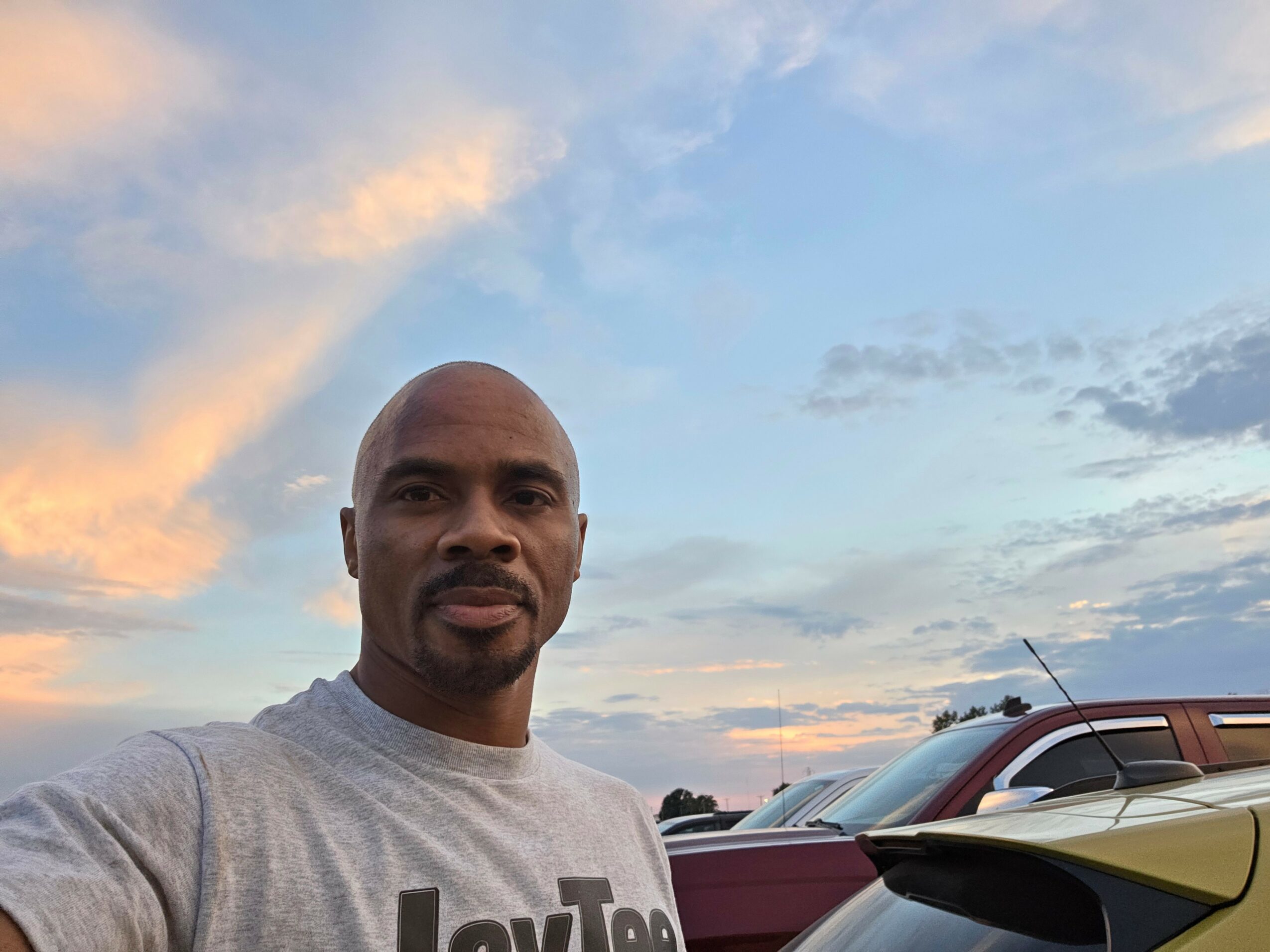 Man with a shaved head and goatee wearing a gray T-shirt outdoors during sunset, with cars and sky in background.