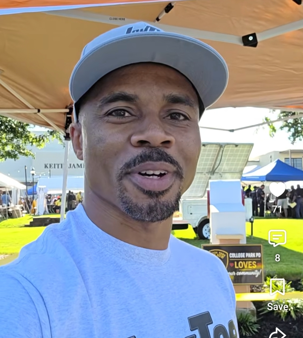 Man wearing a light blue shirt and cap, smiling outdoors under a tent with booths and trees in background.