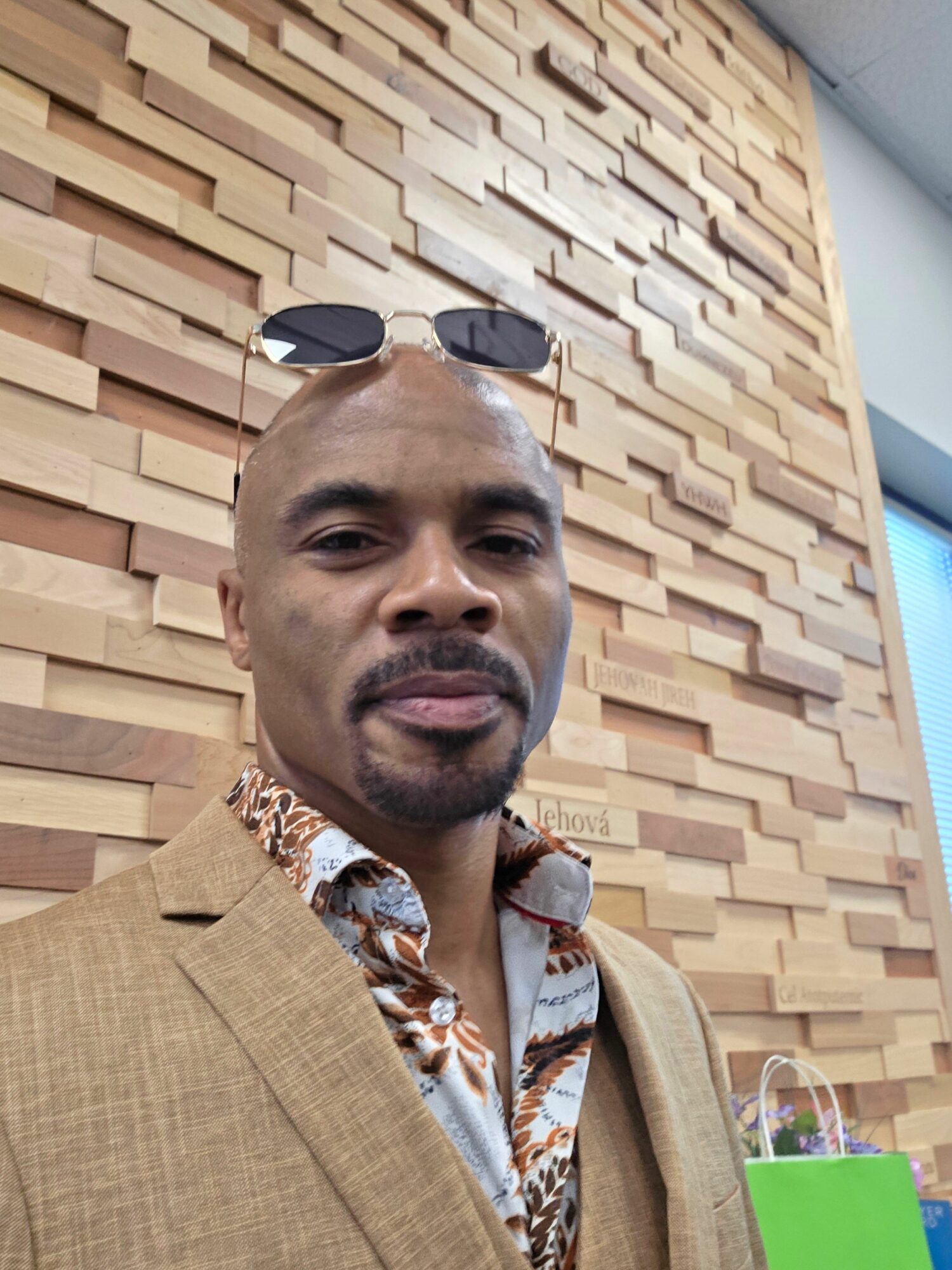 Man with sunglasses and a patterned shirt, sitting against a wooden wall, looking at the camera.