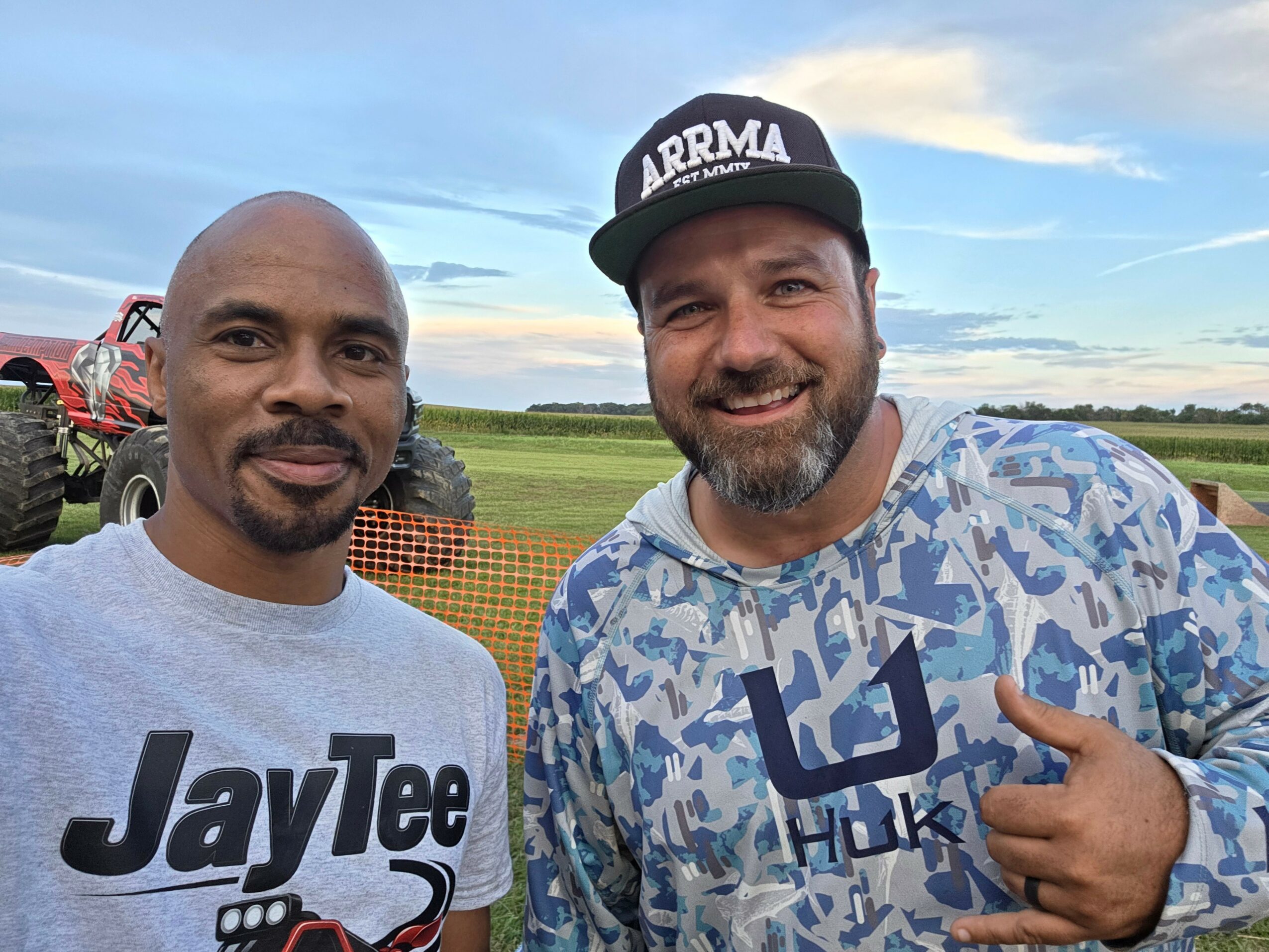 Two men smiling outdoors, one wearing a JayTee shirt and the other in a patterned shirt and cap, pointing at each other.