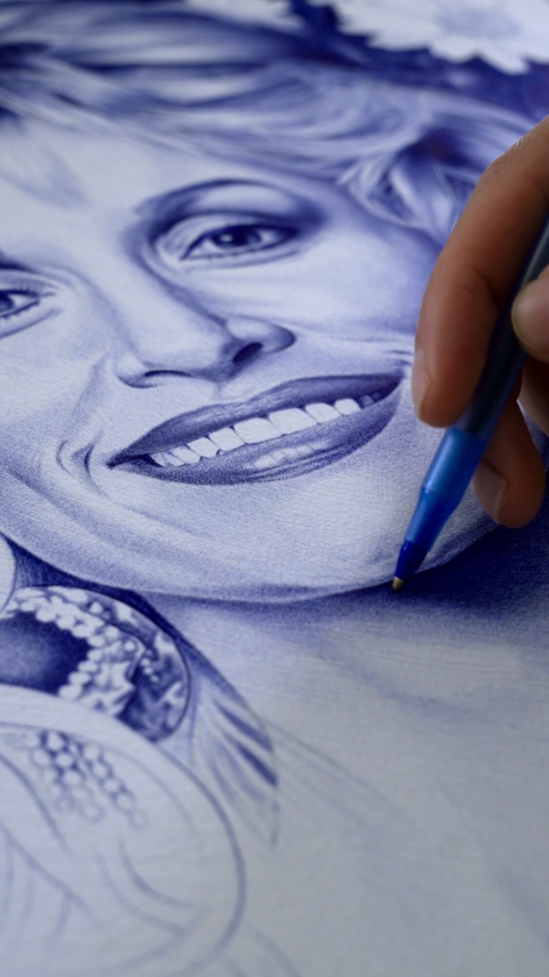 Person's hand drawing a detailed portrait of a smiling woman with jewelry, using a blue pencil.