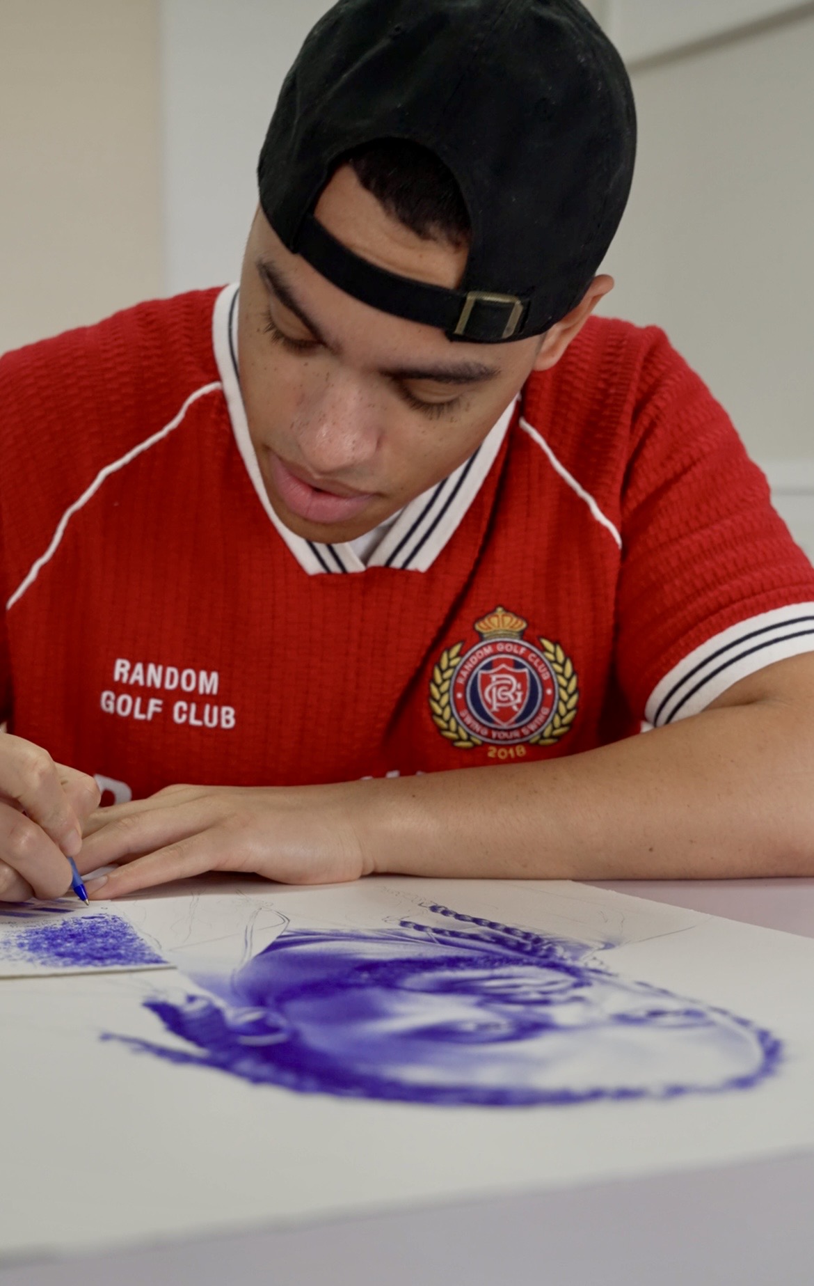 Young person wearing a red shirt and black cap, drawing with purple paint on paper, focused on artwork.