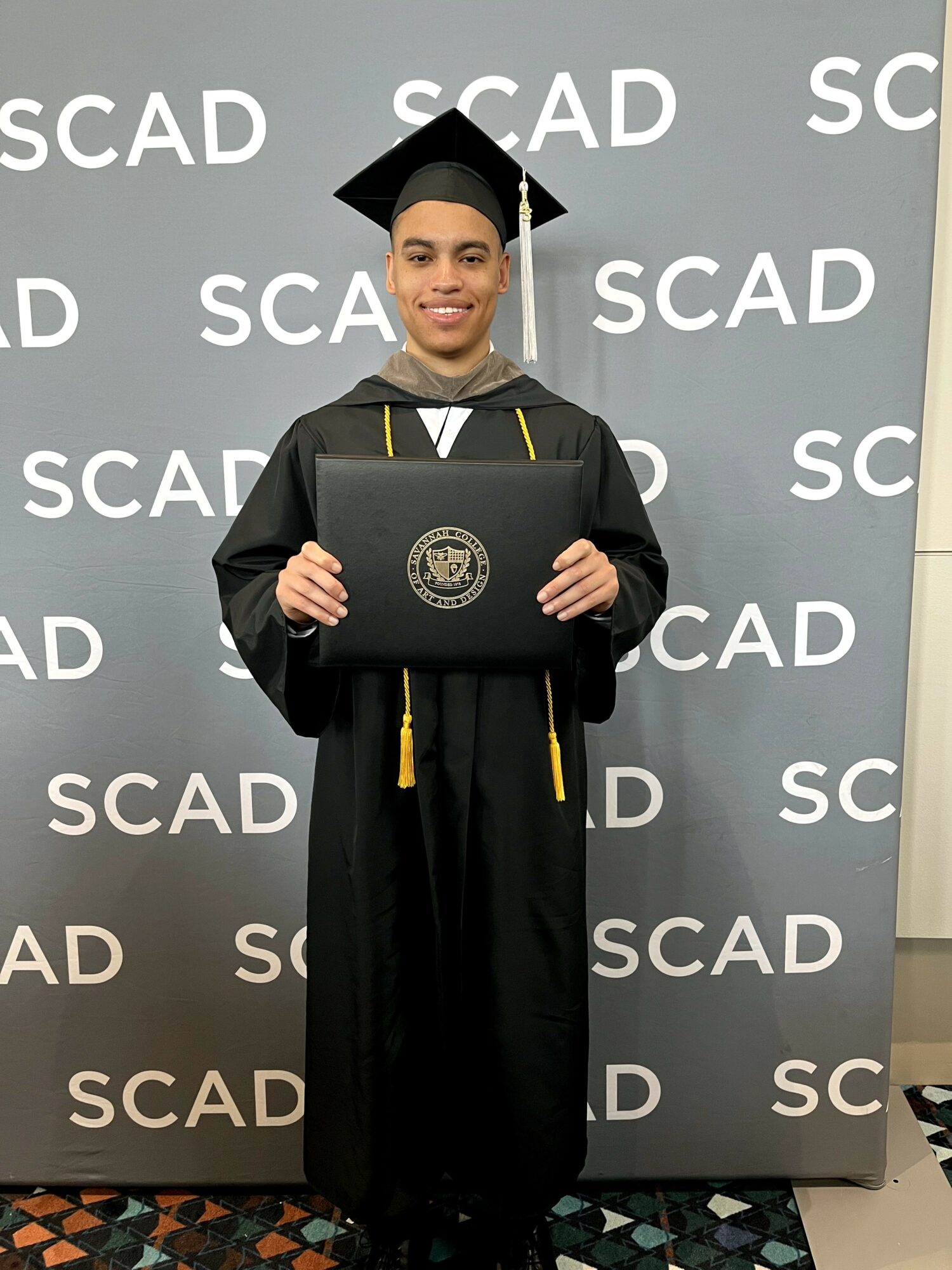 Graduate in cap and gown holding diploma, standing in front of SCAD backdrop, smiling.