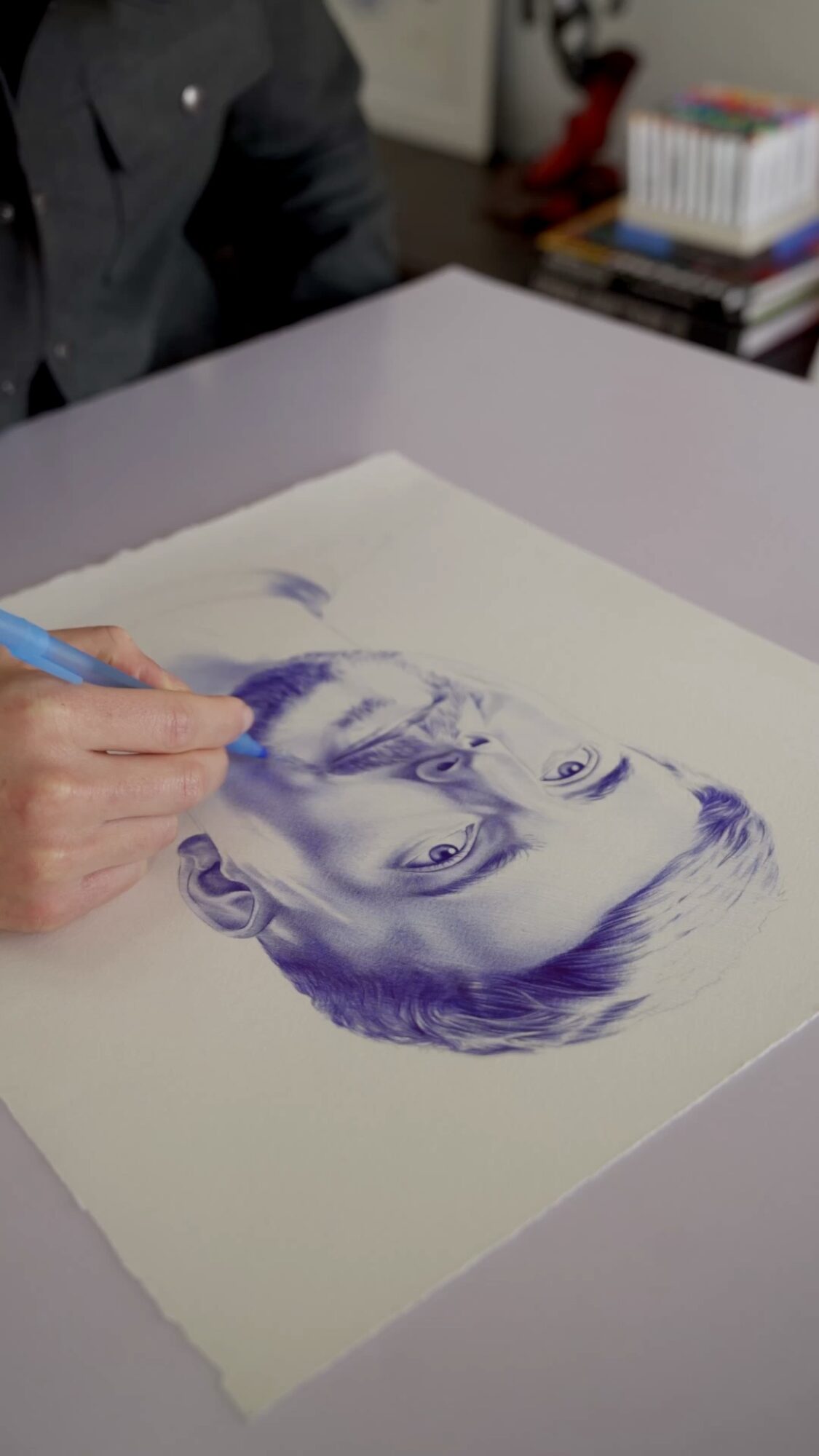 Person drawing a detailed portrait of a woman on paper with a blue pen, on a table with books nearby.