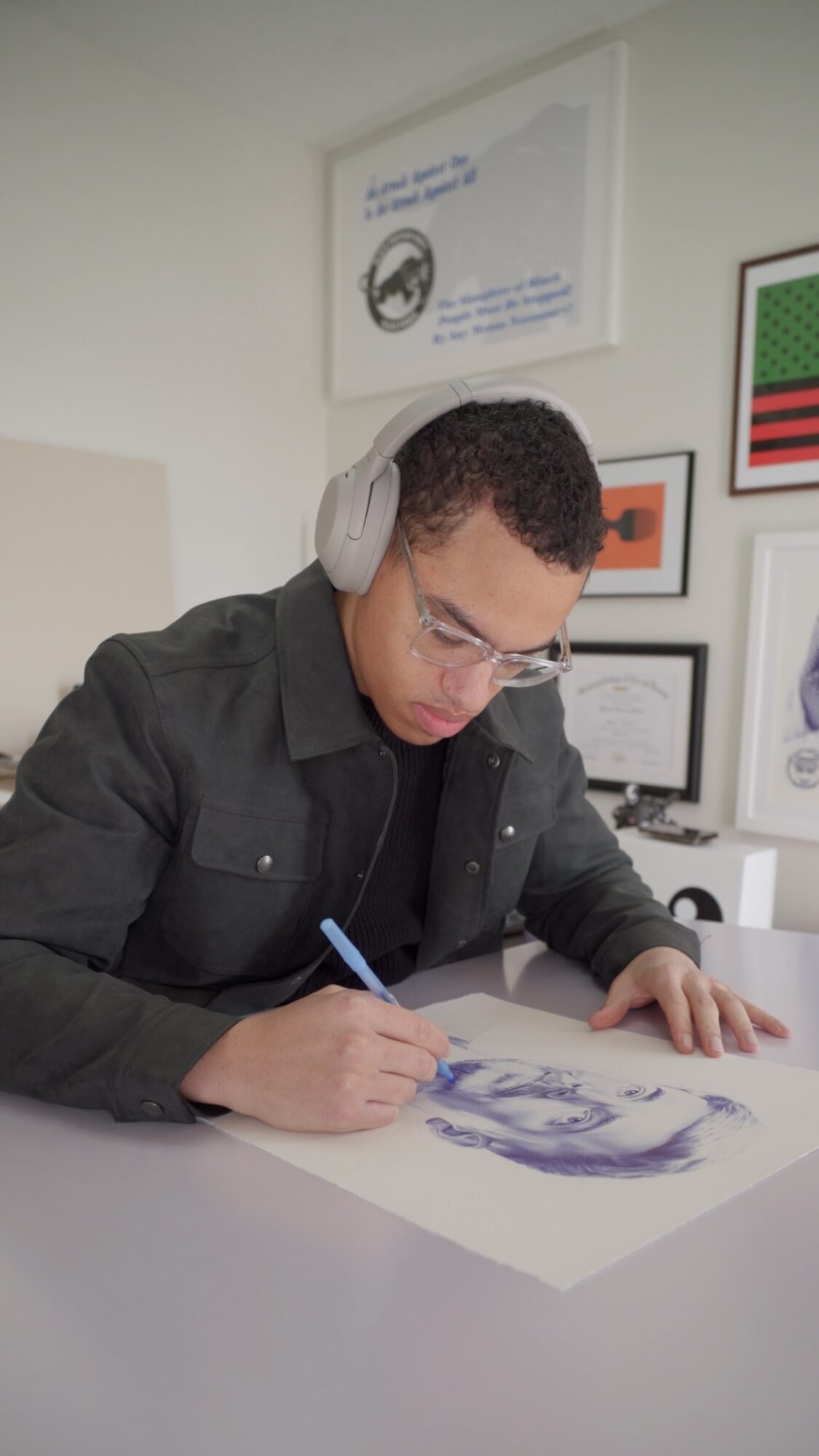 Young man with headphones drawing on paper at desk, surrounded by framed artwork and posters on wall