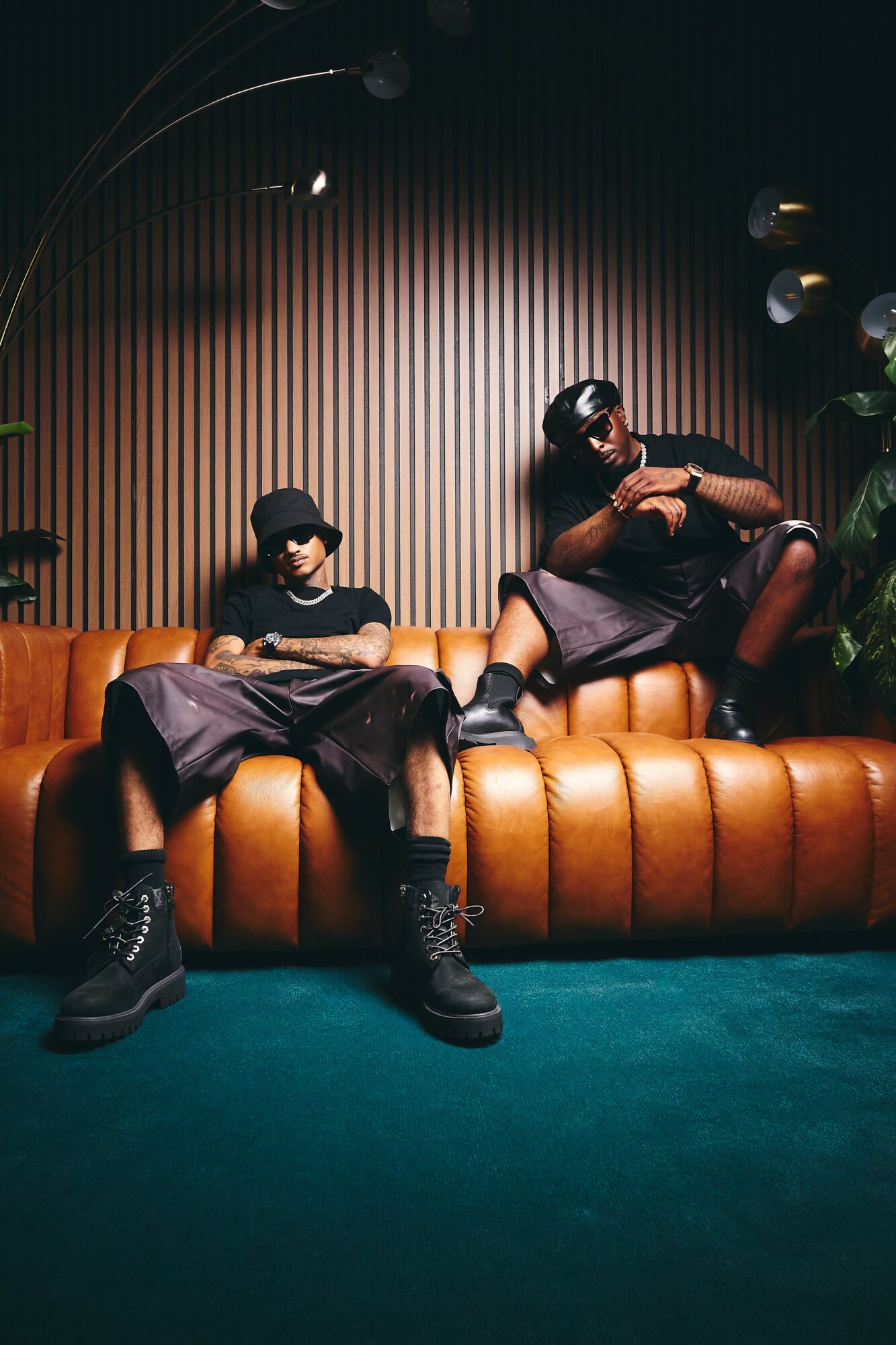 Two people sit on a brown leather sofa in a dimly lit room with a wooden wall background and plants.