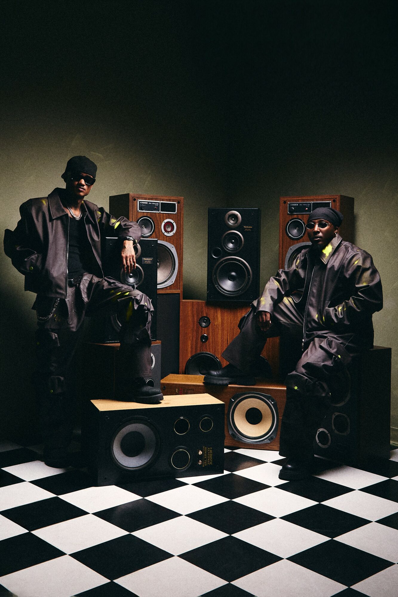 Two people in dark clothing and sunglasses sit among large speakers on a checkered floor, dark background, dim lighting.