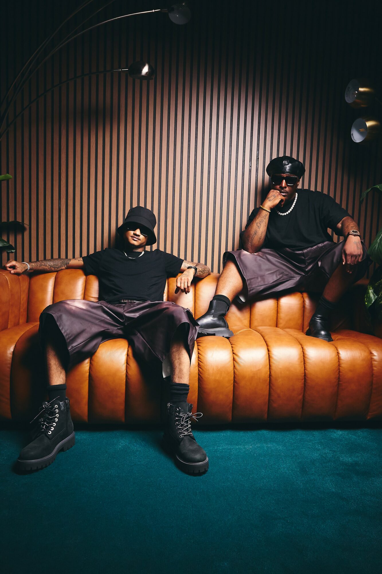 Two people sit on a brown leather couch in a dimly lit room with vertical wooden paneling background.