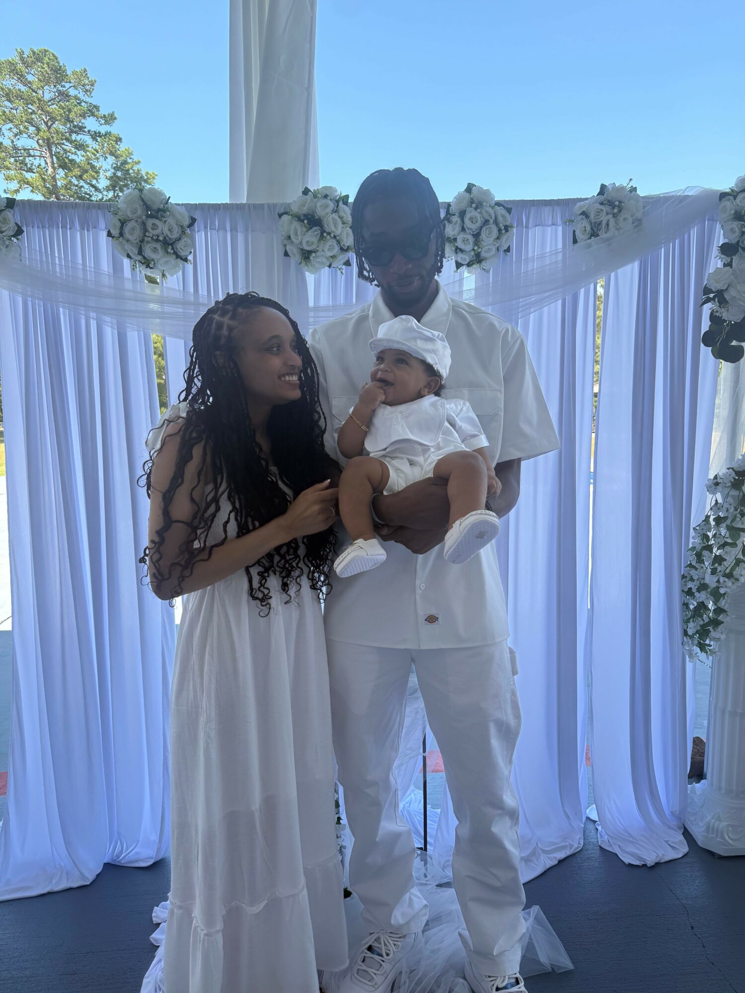 Two people, one in a white suit and the other in a white dress, holding a baby in a decorated setting.