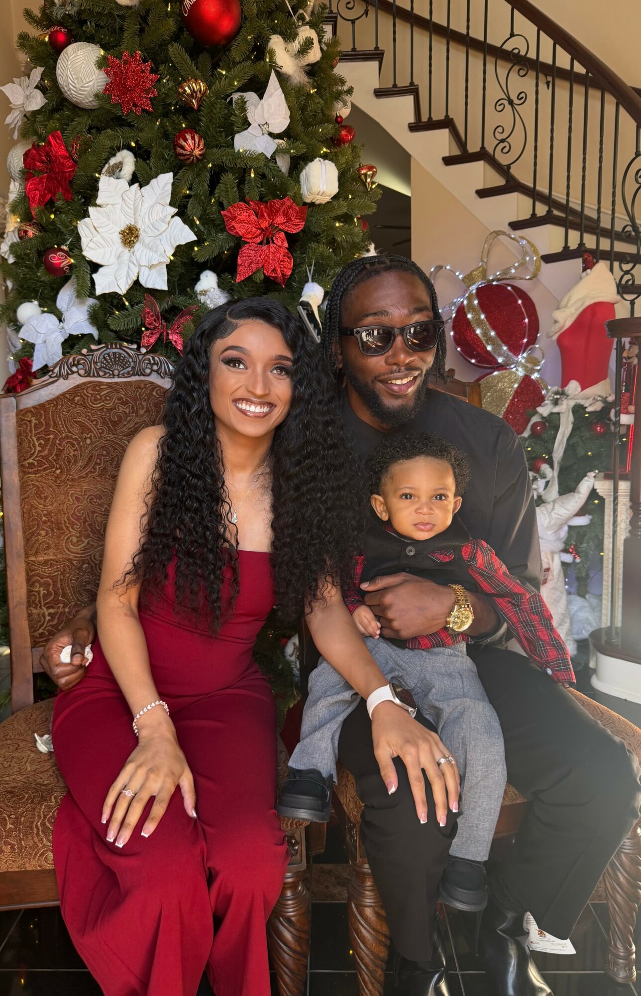 Family sitting in front of Christmas tree, woman in red dress, man with sunglasses, child on man's lap, festive decorations