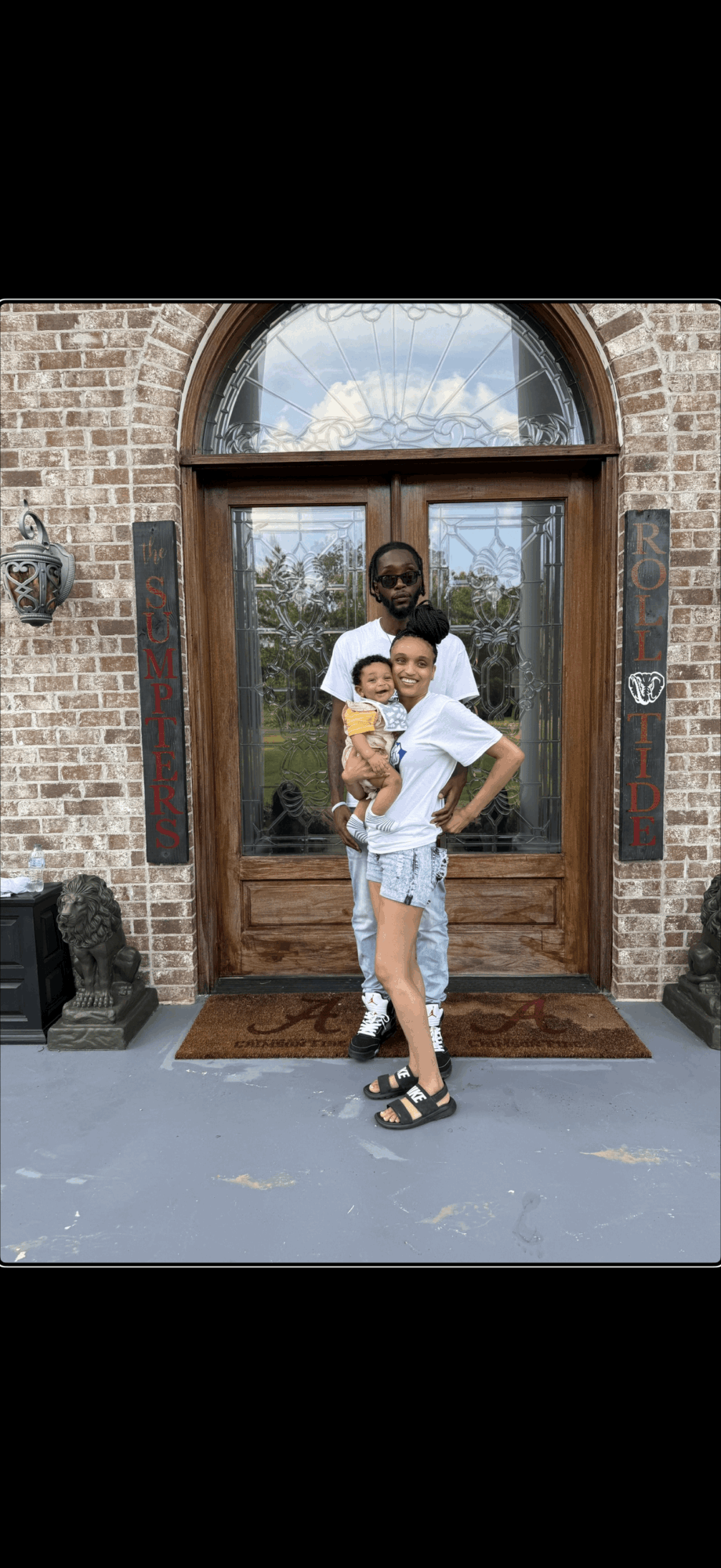 Person holding a child in front of a wooden door with brick wall, glass window, and decorative lanterns, outdoors.