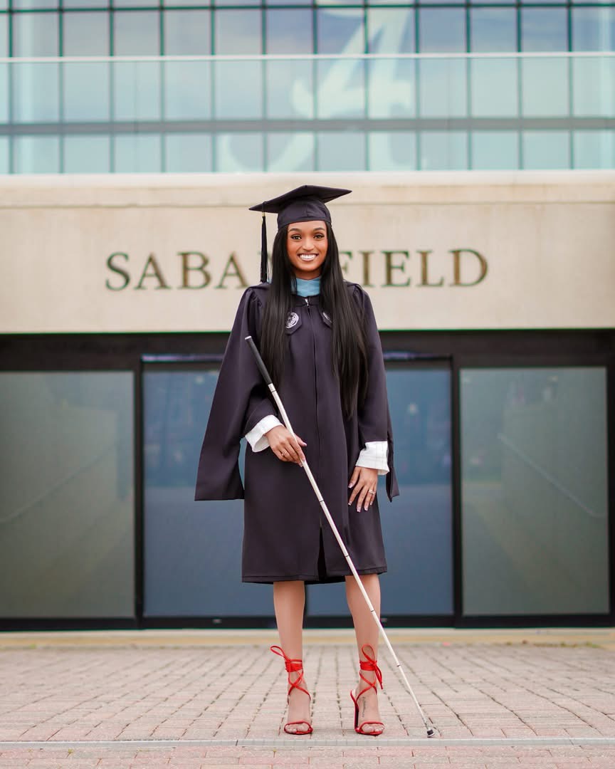 Graduate in cap and gown holding diploma, standing outside building with 'SABA FIELD' sign, smiling, wearing red high heels.
