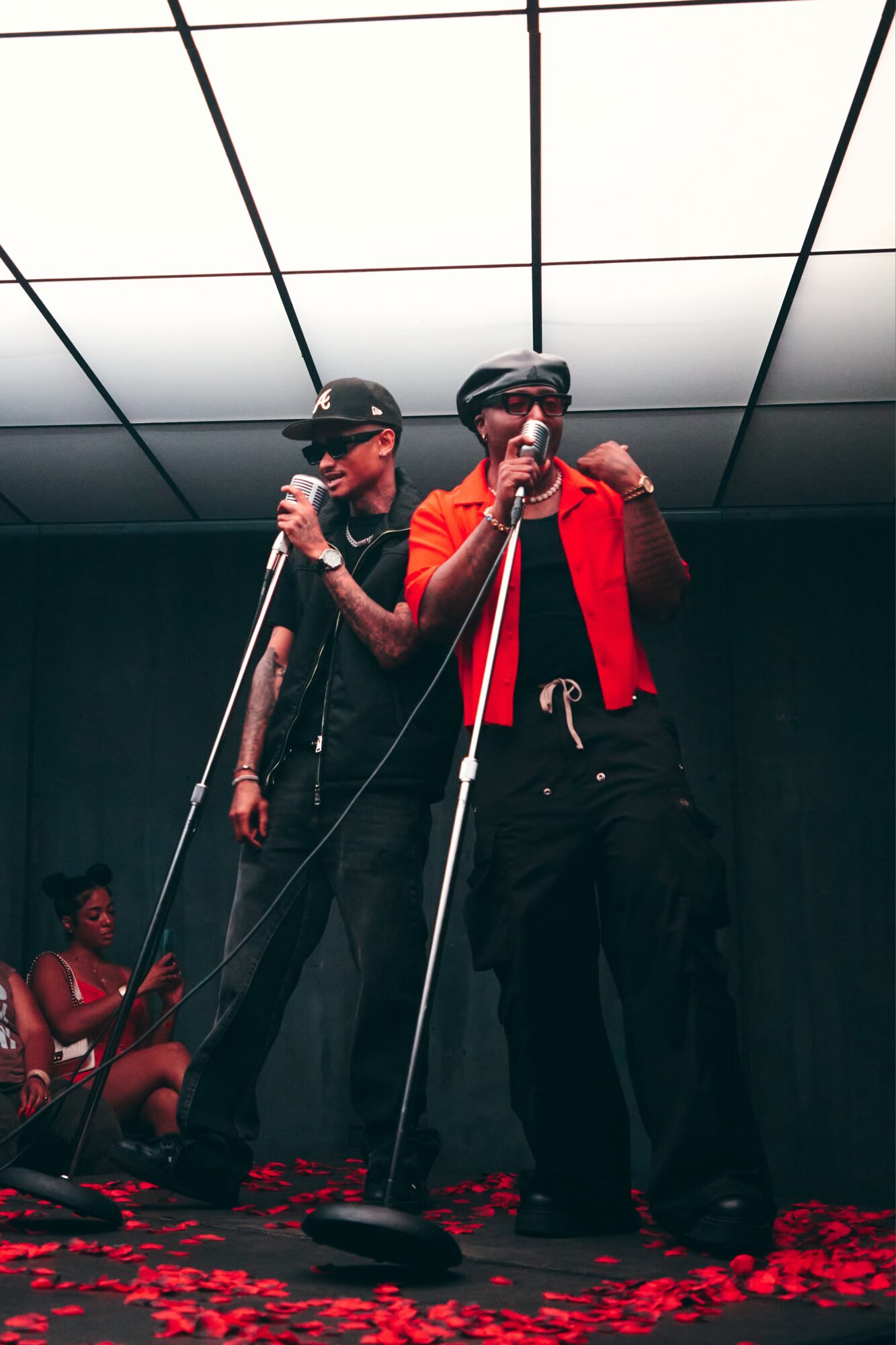 Two performers on stage with microphones, one wearing a red jacket, black pants, and sunglasses, the other in black, with a woman seated in the background.