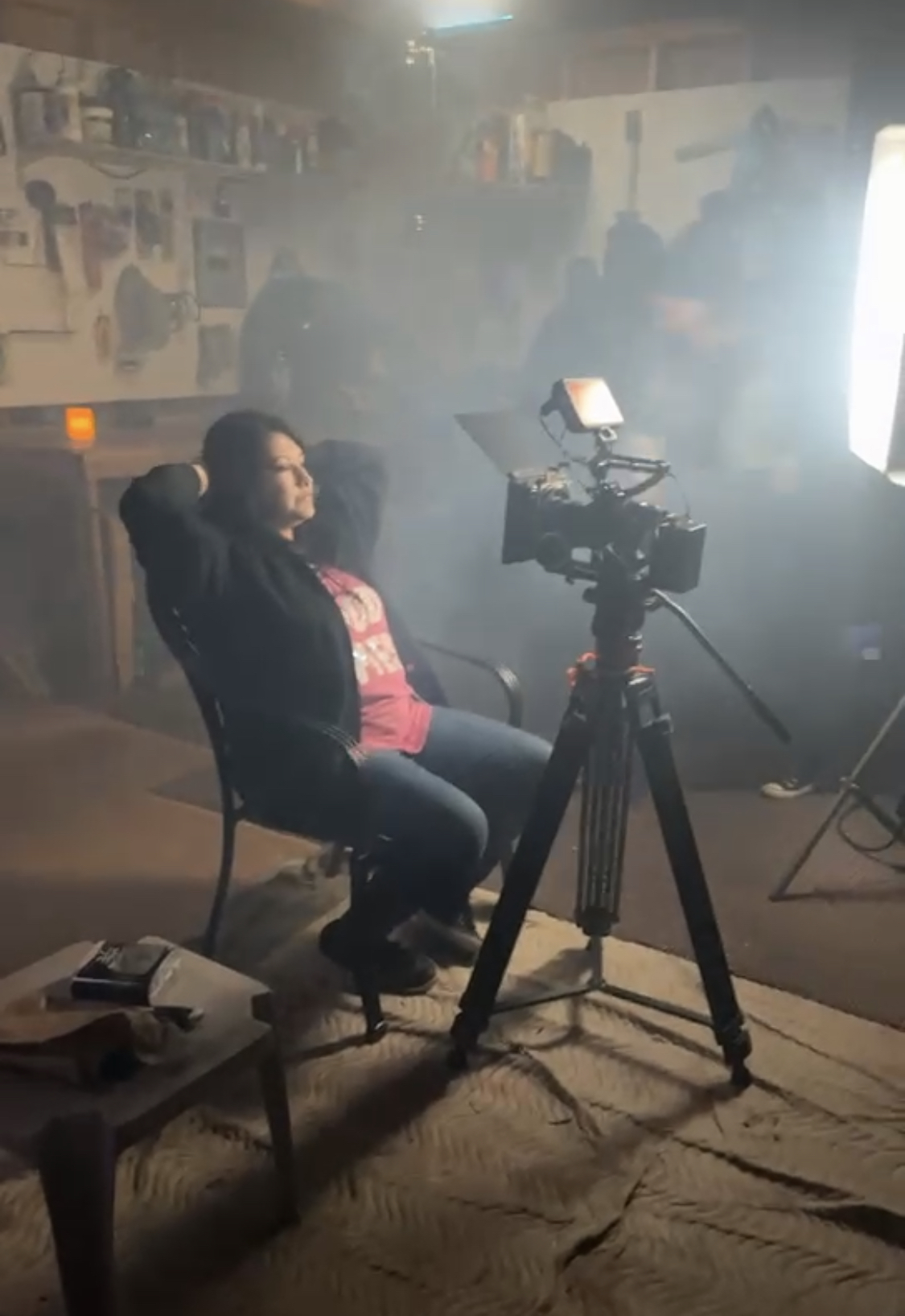 Person sitting in a chair in a room with filming equipment, smoke or fog, and people in the background.