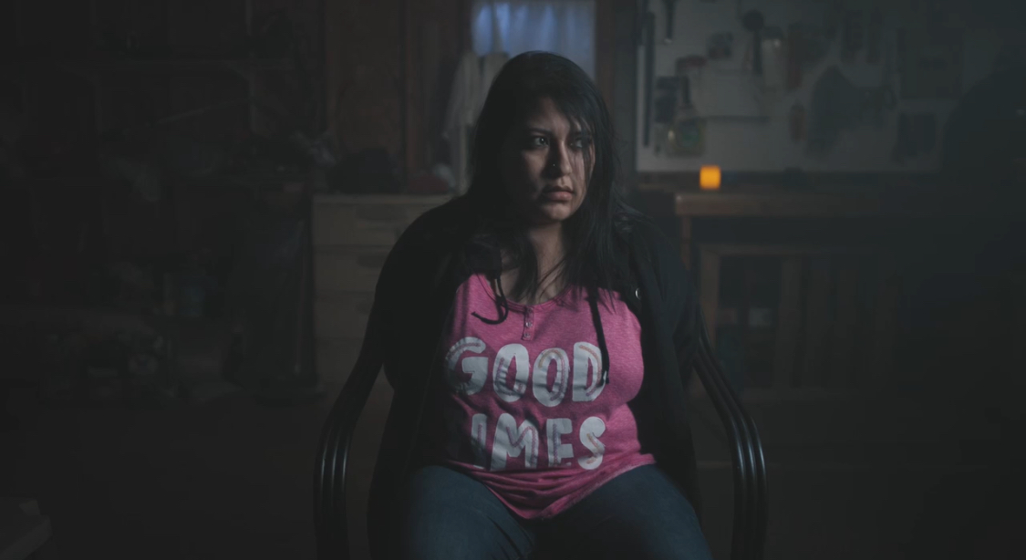 Woman with dark hair sitting in a dimly lit room, wearing a pink shirt with text, looking serious.