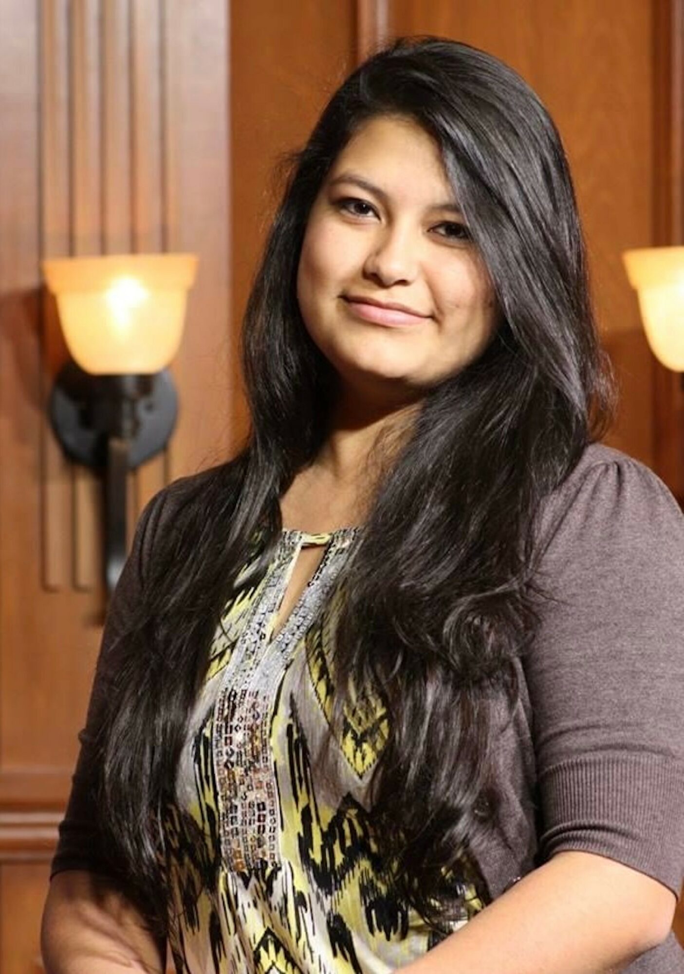 Young woman with long dark hair smiling, standing indoors with wooden paneling and wall sconces behind her.