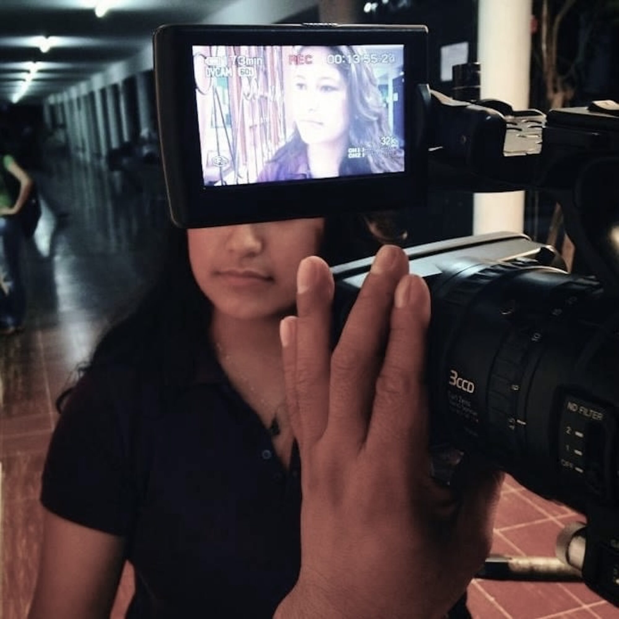Person being filmed by a camera, with the camera's screen showing their face, in an indoor setting.