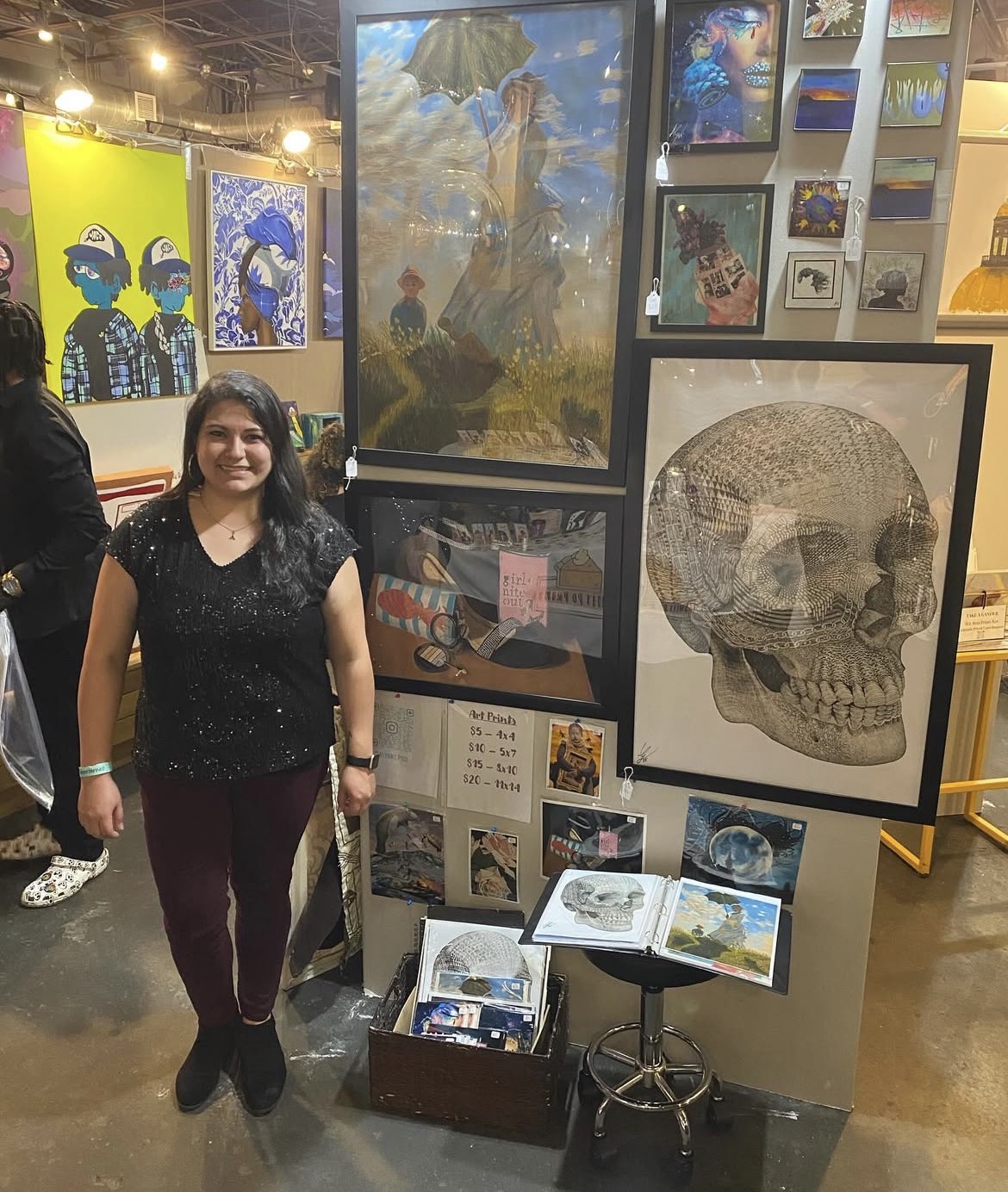 Young woman standing in art gallery with paintings and skull illustration behind her, smiling, holding a bag, in a black top and maroon pants.