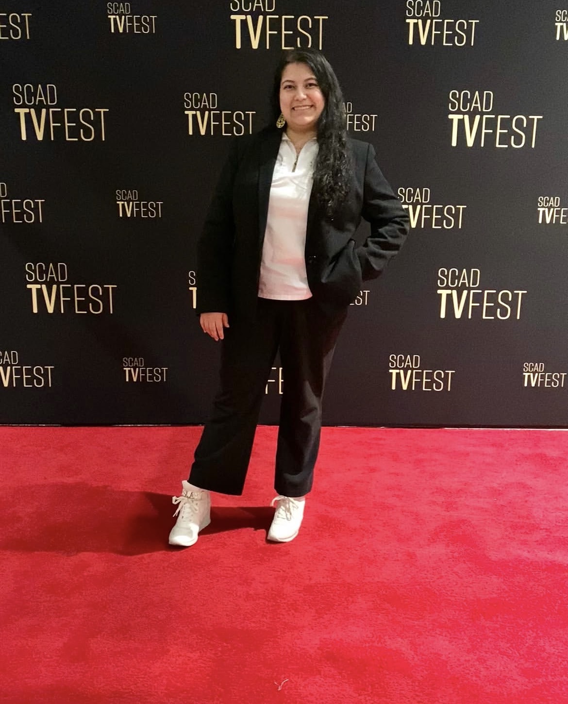Woman standing on red carpet in front of black backdrop with TV festival logos, smiling, wearing black jacket, white shirt, black pants, white shoes.