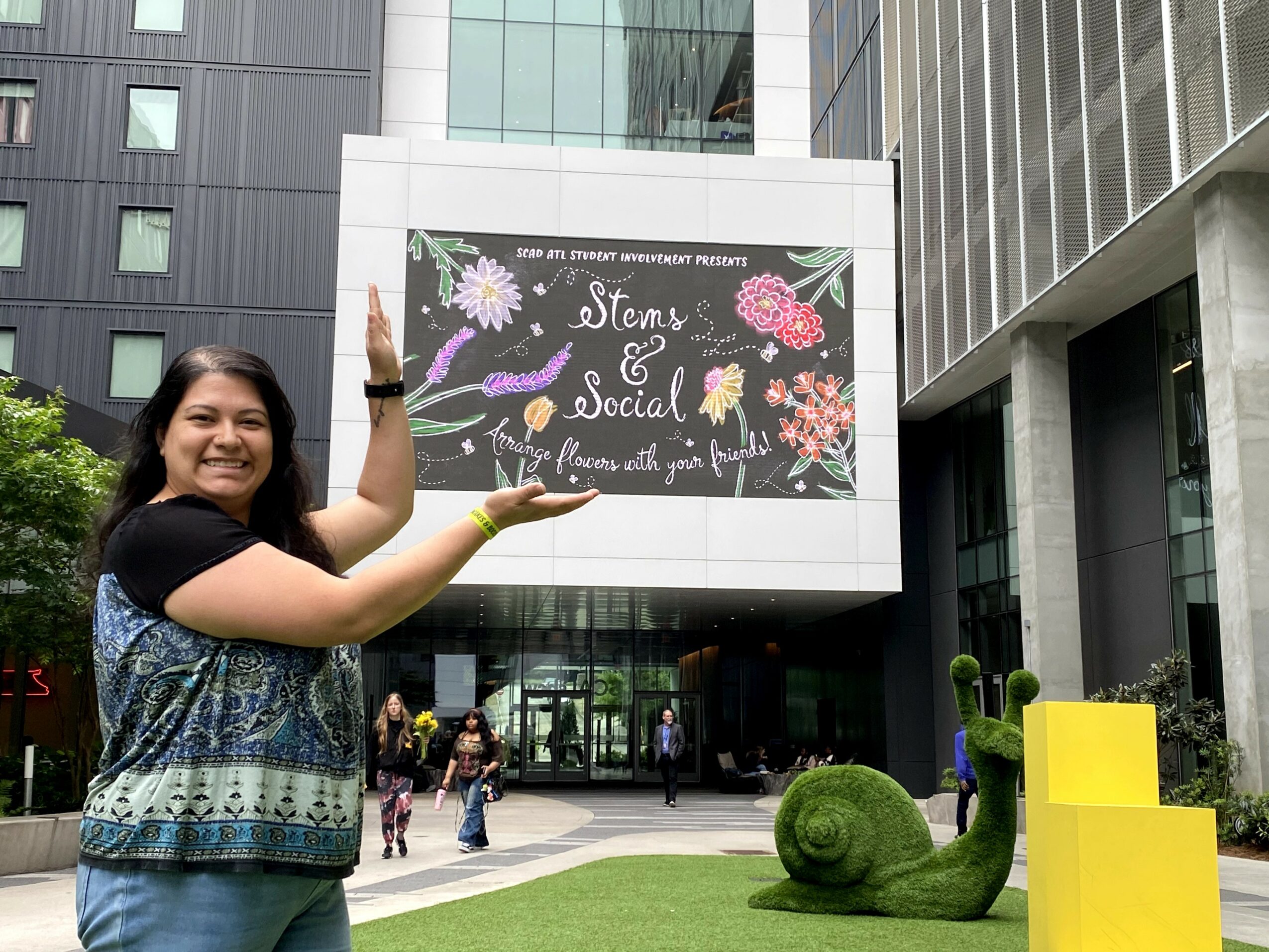 Smiling woman standing outside a modern building with a large sign reading 'Stay & Social' and floral decorations, people walking nearby.