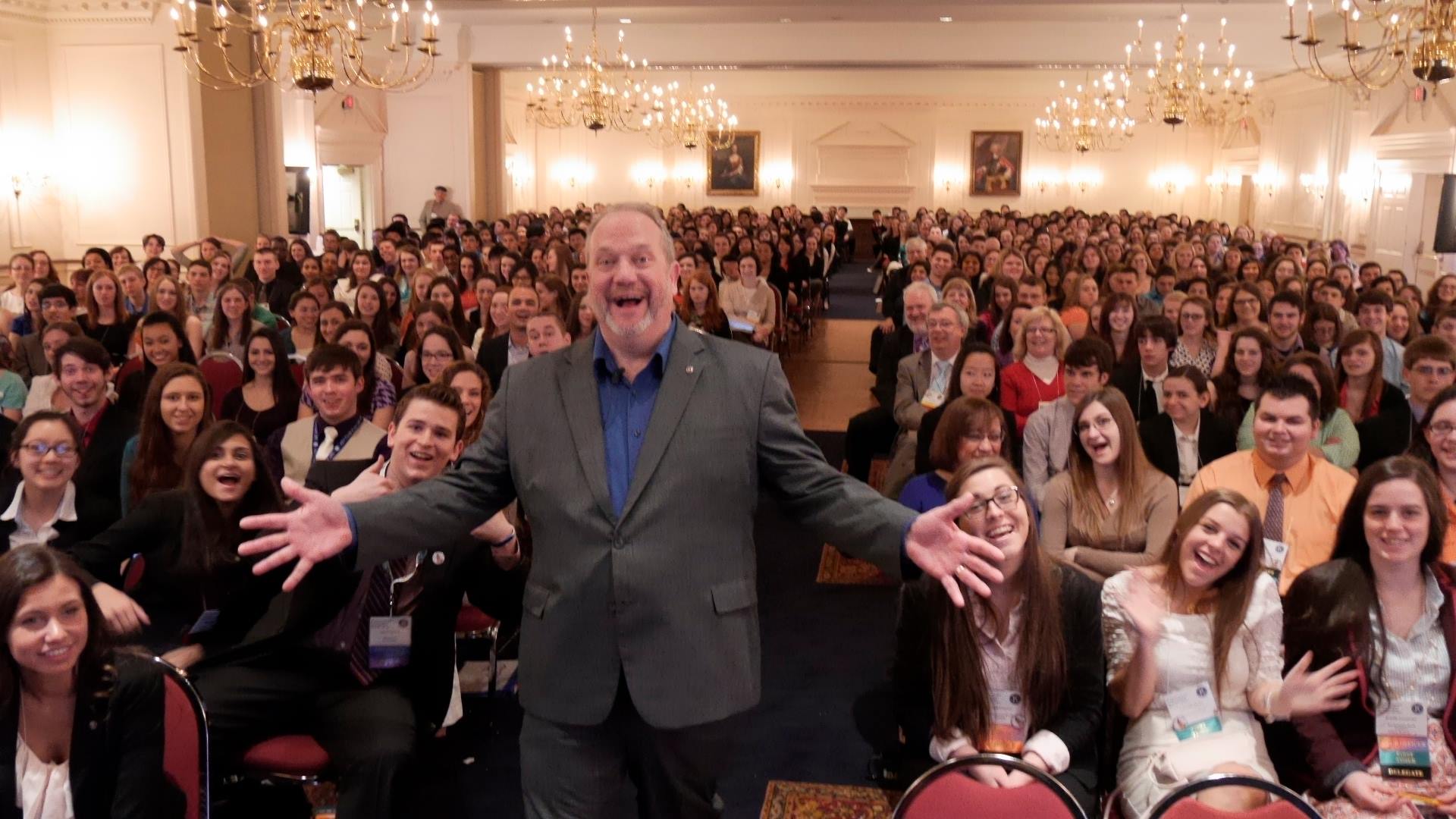 A man in a suit stands with arms outstretched in front of a large audience in a banquet hall, smiling.