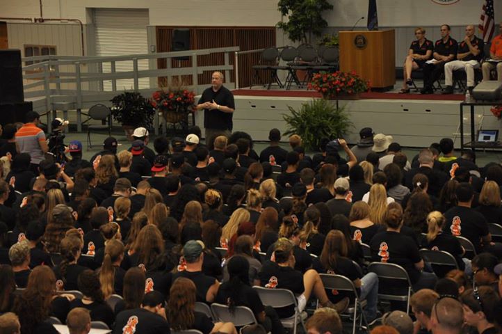 Person speaking to large audience seated indoors, with panel of people on stage and plants, audience facing forward.