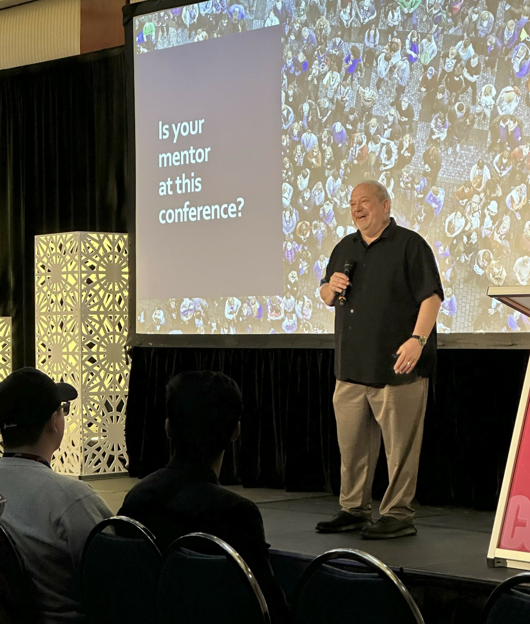 A man stands on stage holding a microphone, with a large screen behind him displaying a question about mentors at a conference.