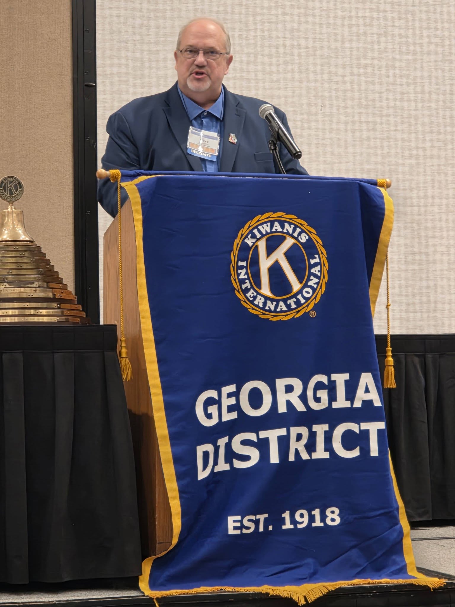 Man standing behind a podium with a blue and yellow Lions Club banner, speaking at an event, in a conference room.