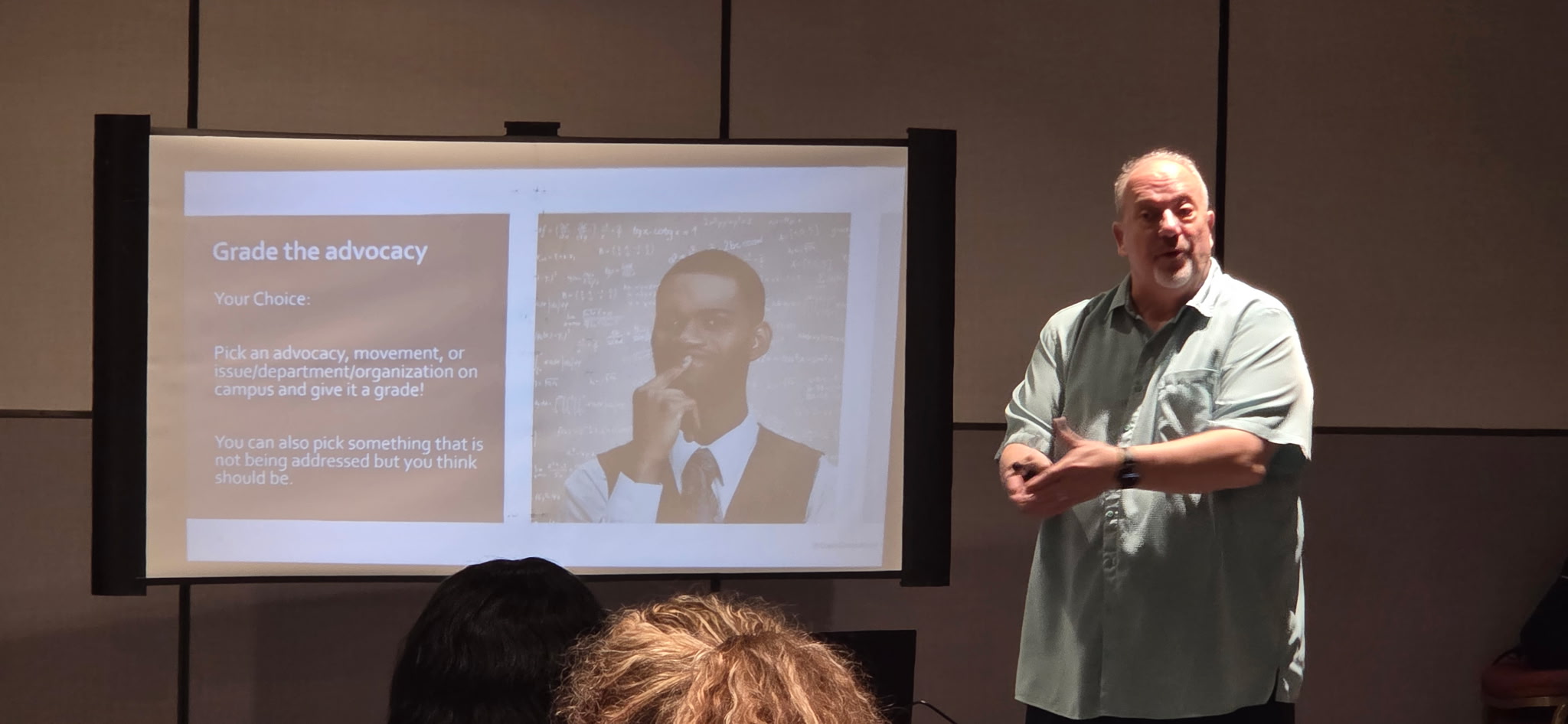 Man giving a presentation in front of a screen with a slide showing a person and text, audience visible in foreground.
