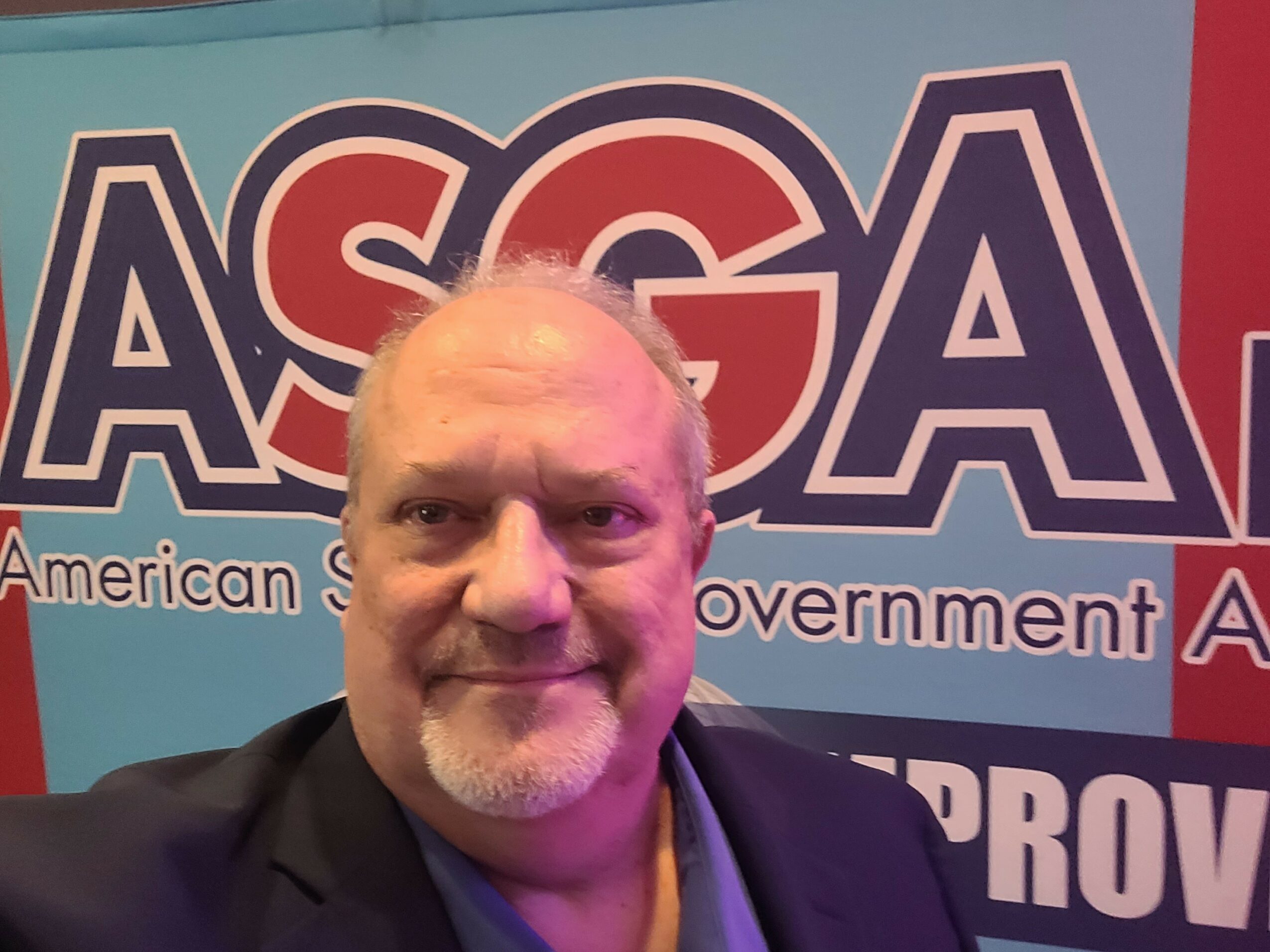 A man with a beard and short hair smiling in front of a blue and red background with the letters 'ASGA'.