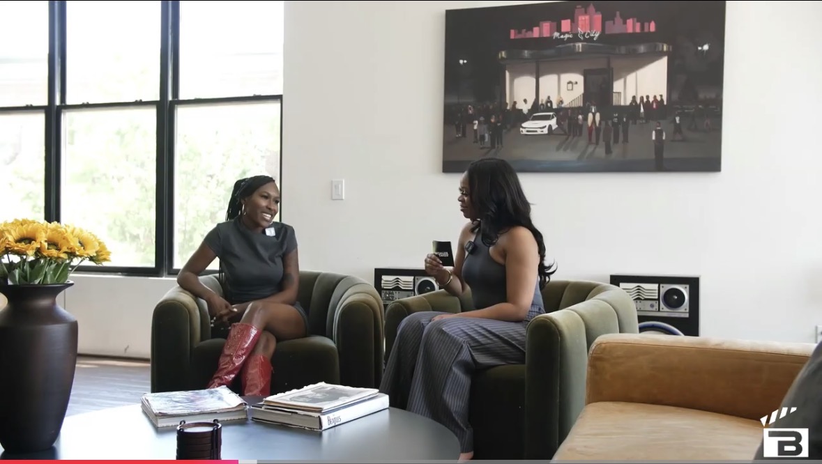 Two women sit on armchairs in a modern room, talking and smiling, with a large window and artwork on the wall.