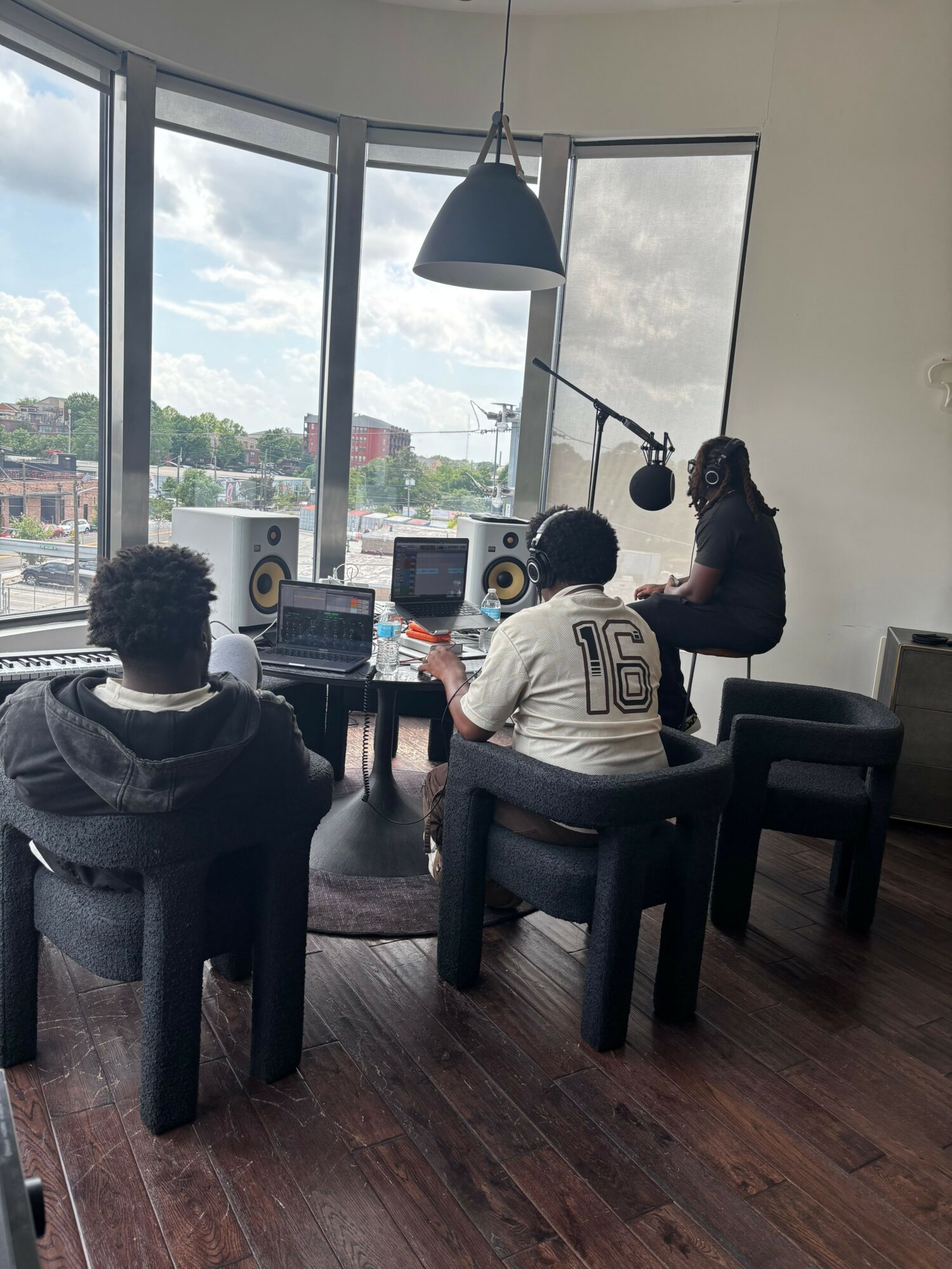 Three people working in a room with large windows, desks, and computers, with a wooden floor and a floor lamp.
