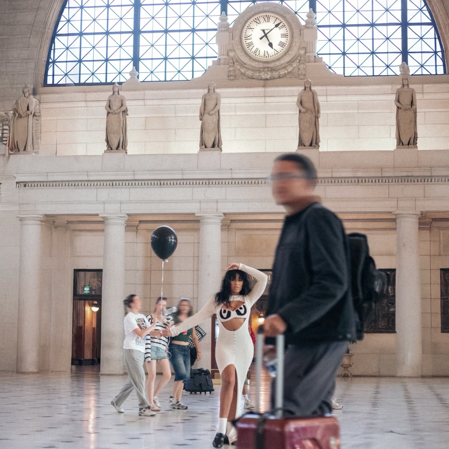 People inside a grand hall with a clock and statues, some walking, one woman in a dress, a man with a suitcase.