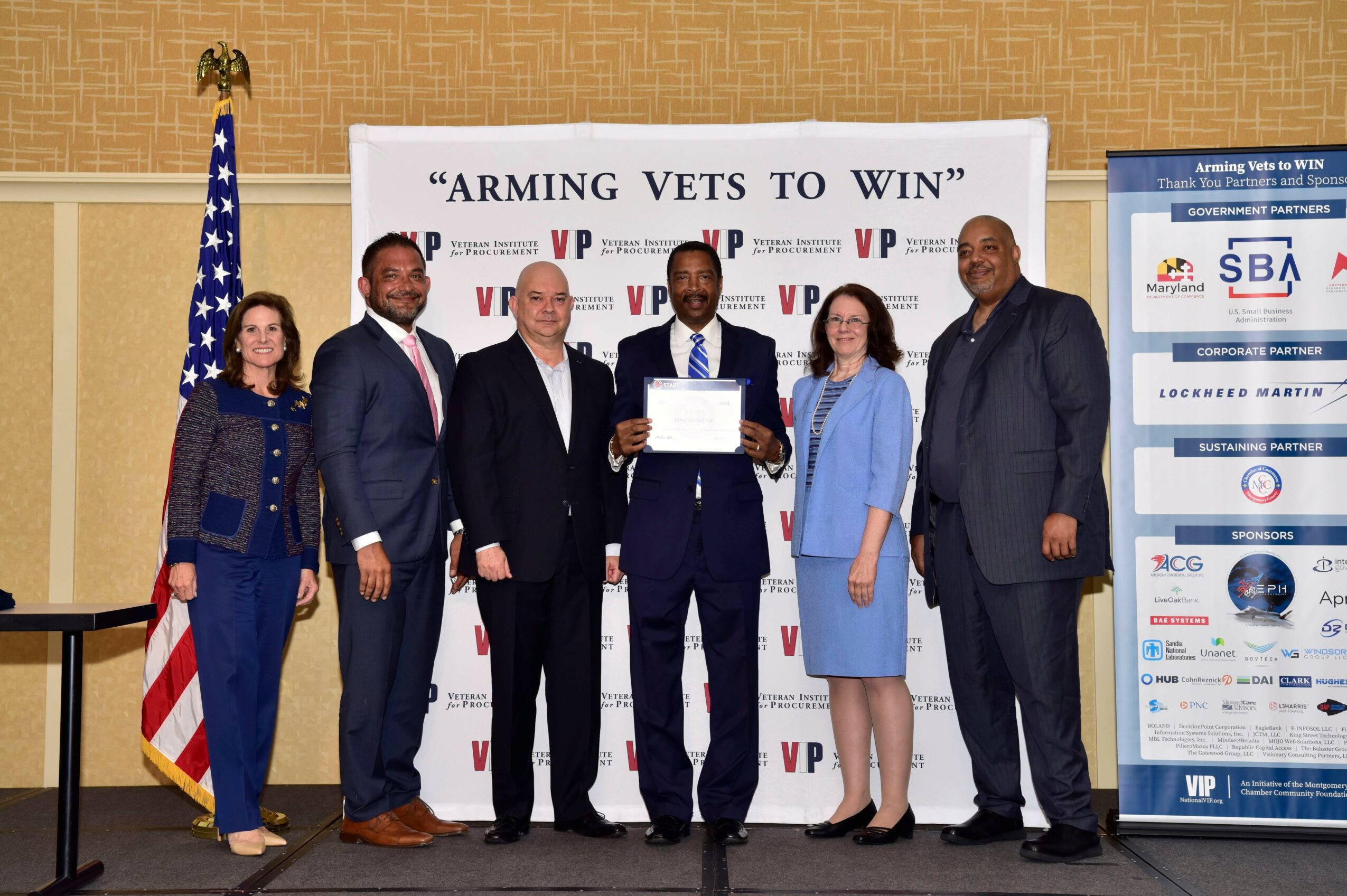 Group of six people standing in front of a banner, with an American flag on the left, at an award ceremony.