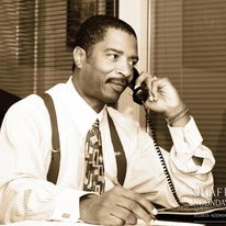 Man in formal attire talking on a landline phone, sitting at a desk with blinds in the background.