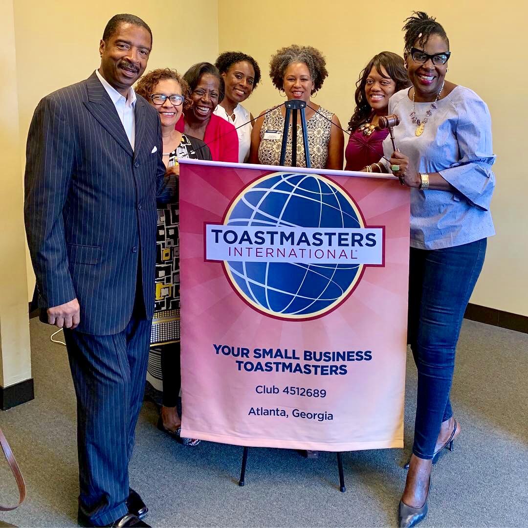 Group of seven diverse people holding a Toastmasters banner, smiling, indoors, in a professional setting.