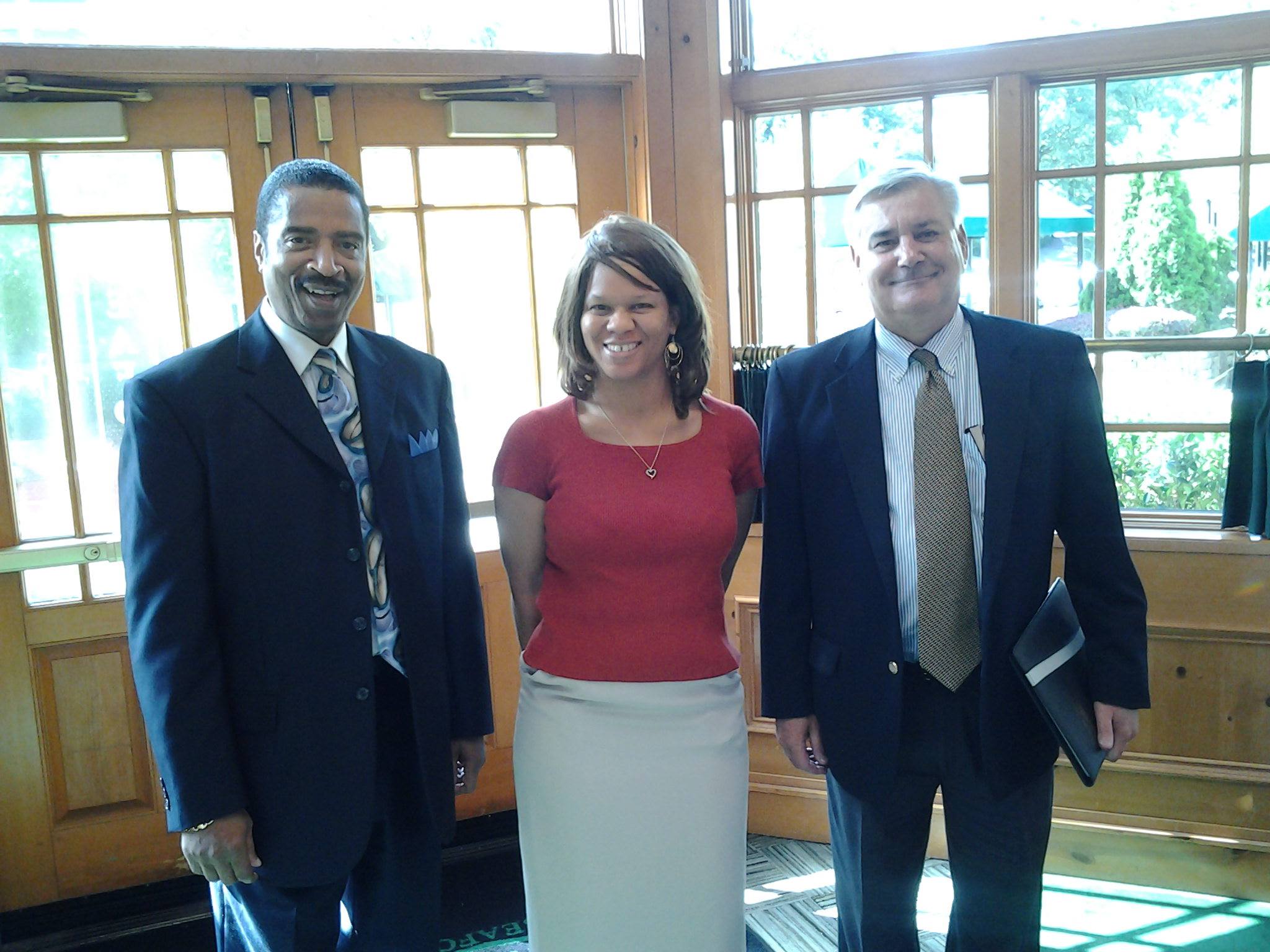 Three people standing indoors near large windows, smiling, dressed in formal attire, with wooden walls and sunlight outside.