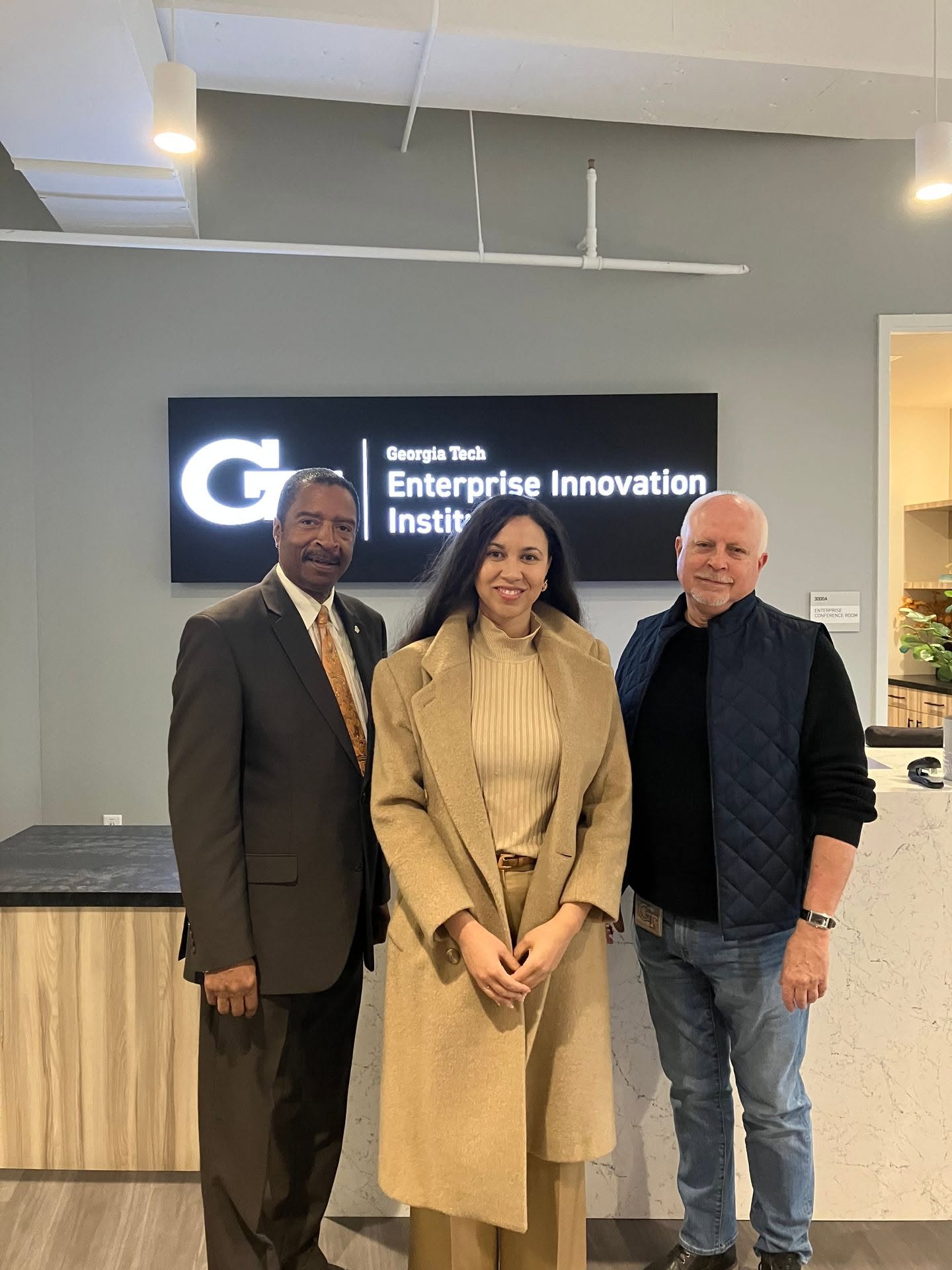 Three people standing indoors in front of a sign for Georgia Tech Enterprise Innovation Institute, smiling.