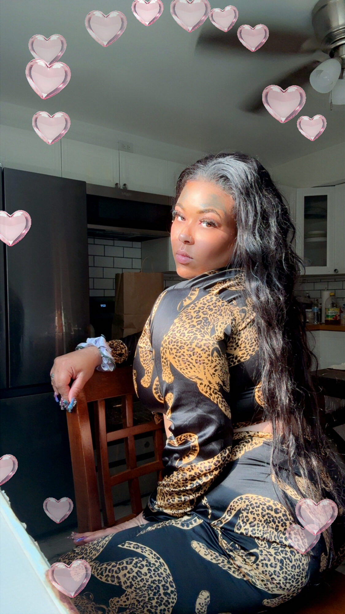 Woman with long wavy hair wearing a patterned outfit, sitting on a chair in a room with kitchen background, pink heart overlay.