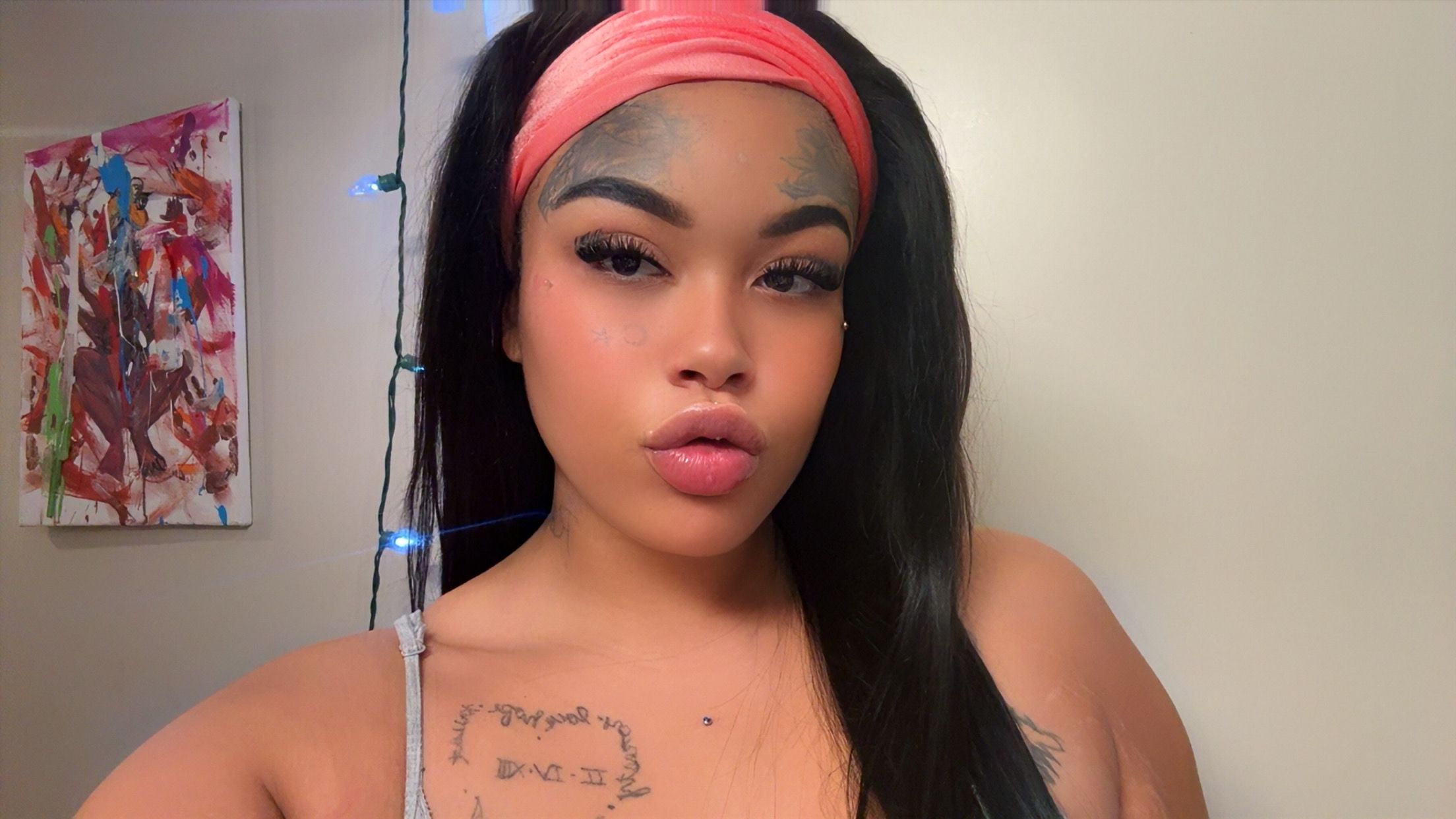 Woman with long dark hair, pink headband, makeup, and a tattoo on her shoulder, in front of a wall with artwork and string lights.