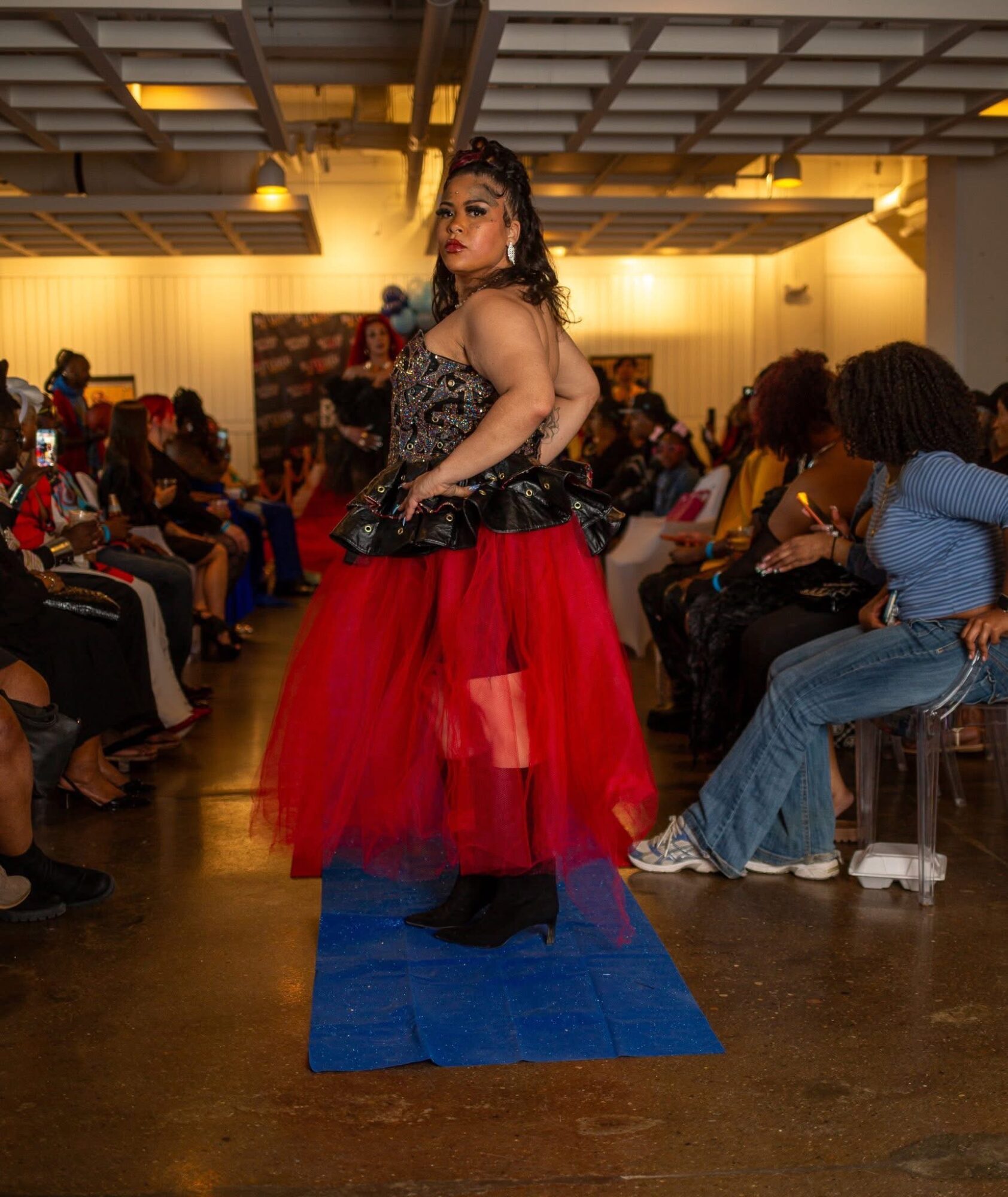 Person walking on a runway with an audience on both sides, wearing a red and black dress, in an indoor setting.