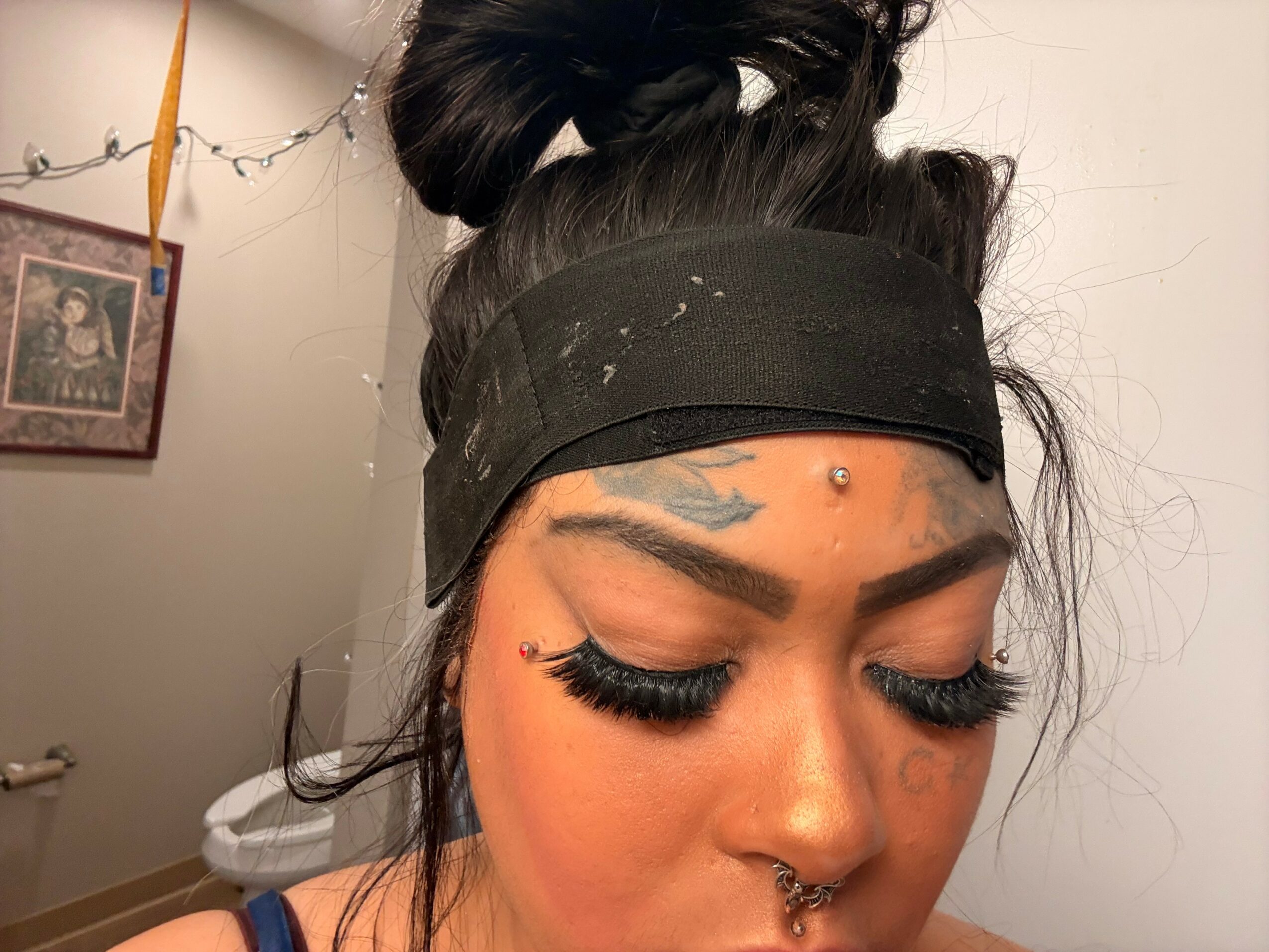 Close-up of a woman with dark hair in a bun, wearing a black headband, with makeup and a septum piercing.