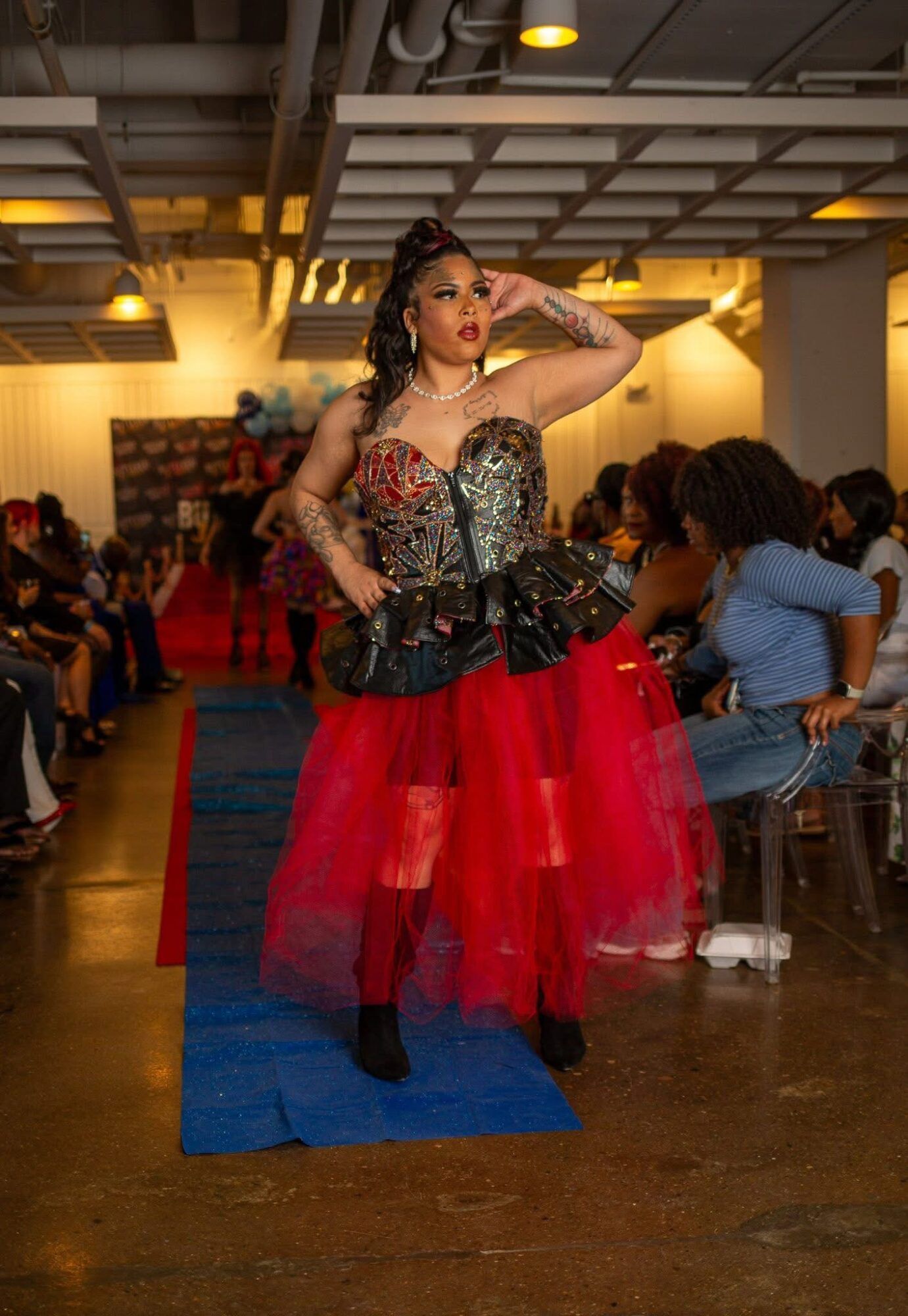 Woman in a red and black dress with a tulle skirt, standing on a blue carpet in a room with seated audience.