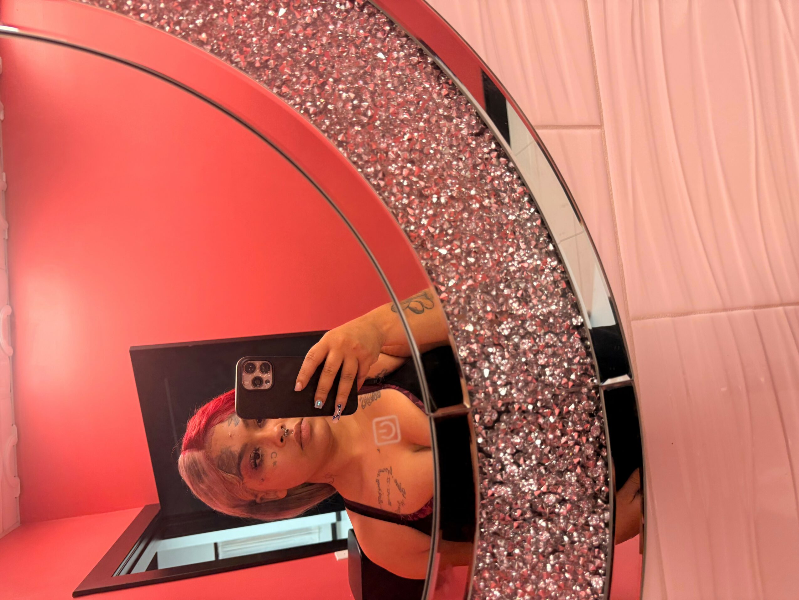 Person taking a mirror selfie with a phone, reflection shows a woman with blonde hair, in a room with pink and glittery decor.