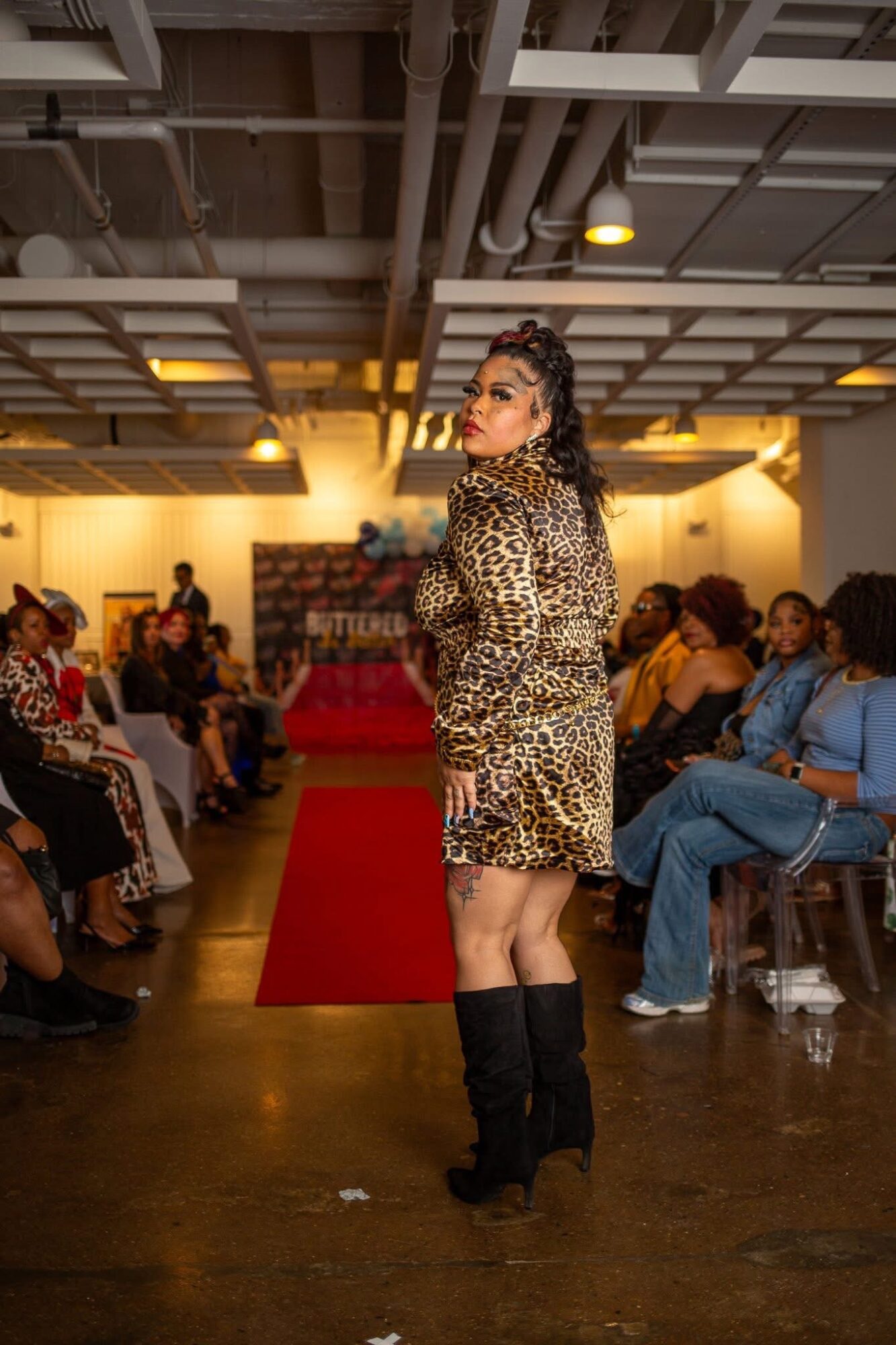 Woman in leopard print dress and black boots standing on a red carpet in a room with seated audience, ceiling lights, and industrial ceiling.