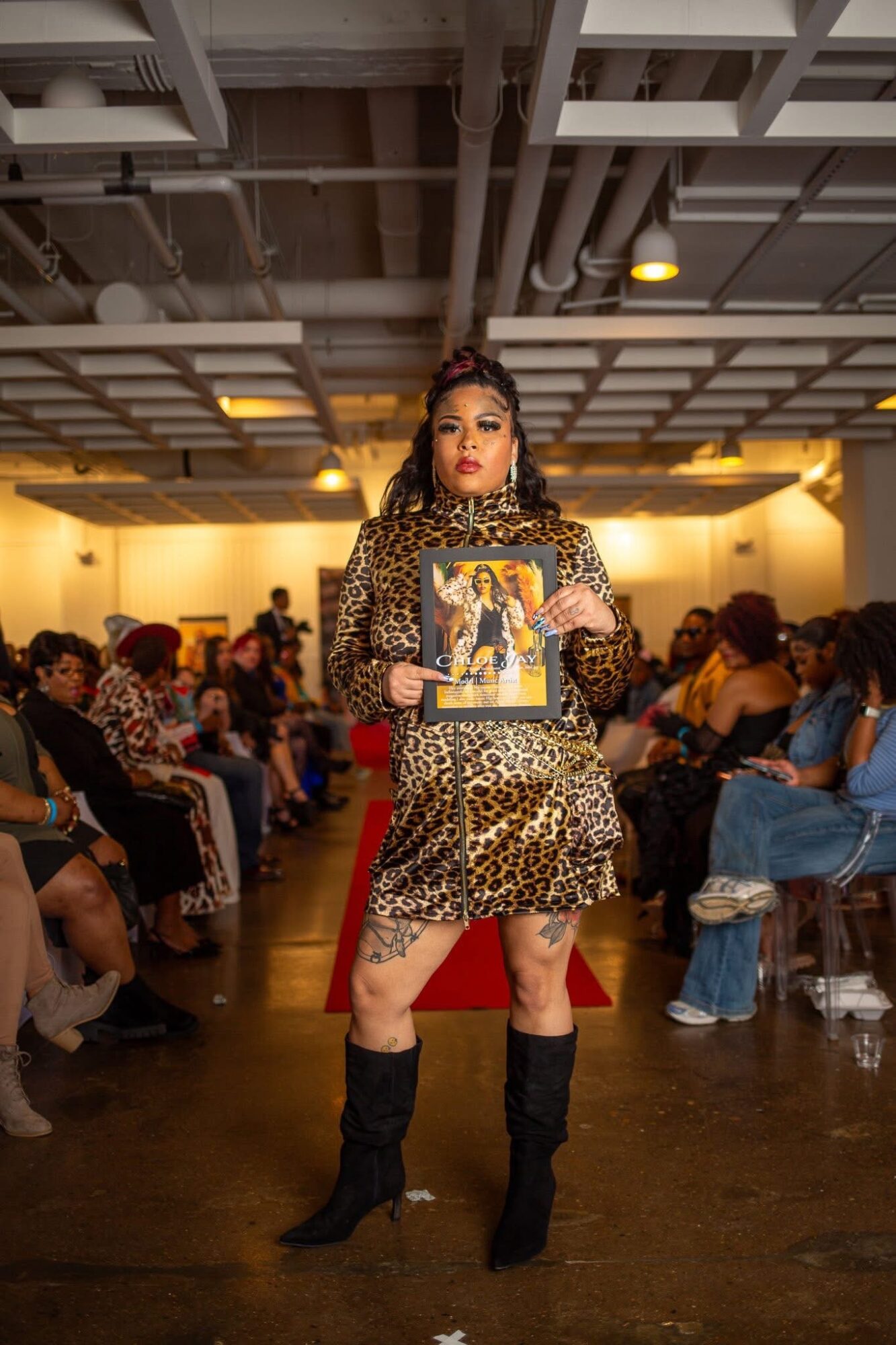 Woman in leopard print dress holding a magazine, standing in a crowded indoor event space with a high ceiling.