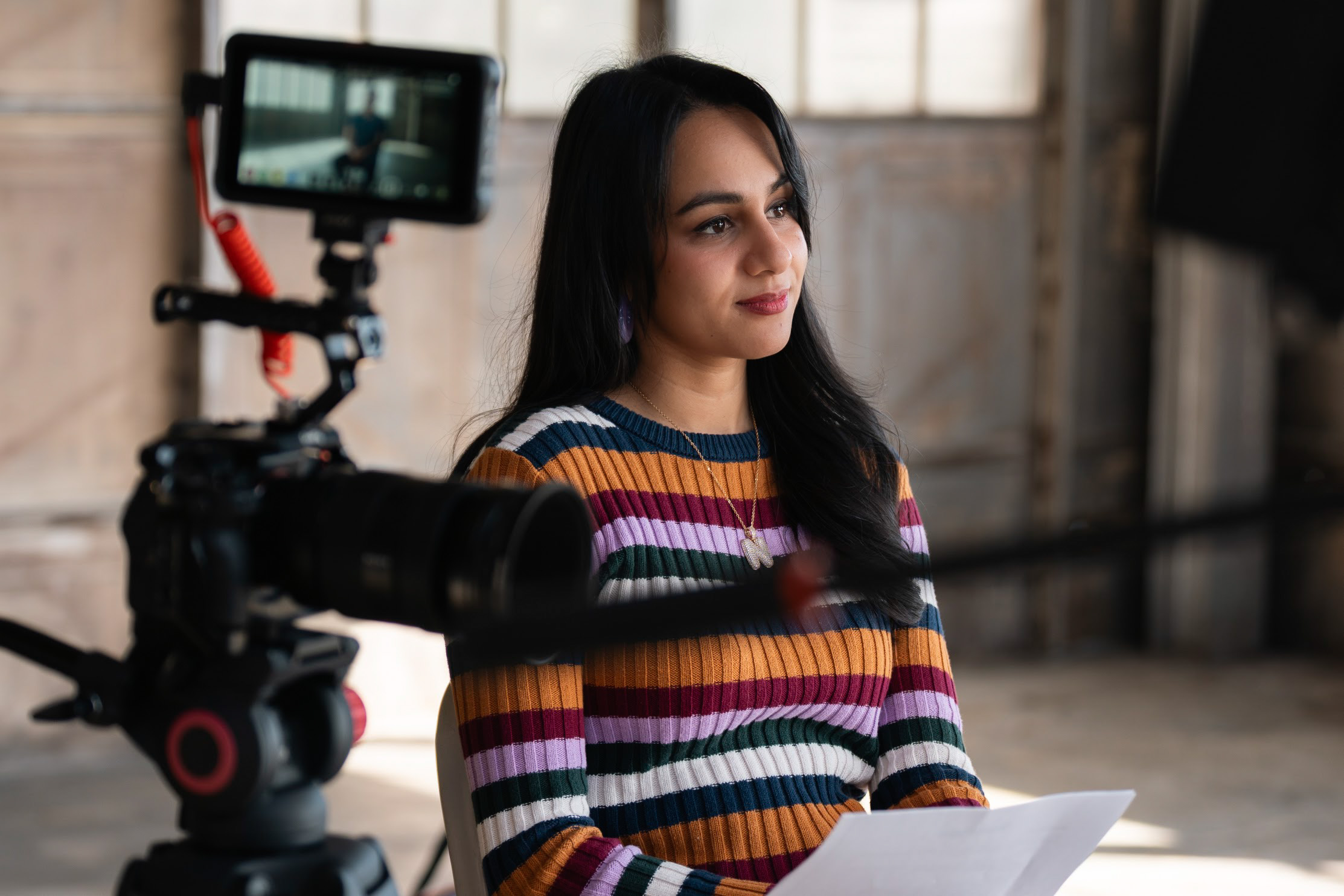 Young woman with long dark hair wearing a colorful striped sweater, sitting indoors with a camera filming her.